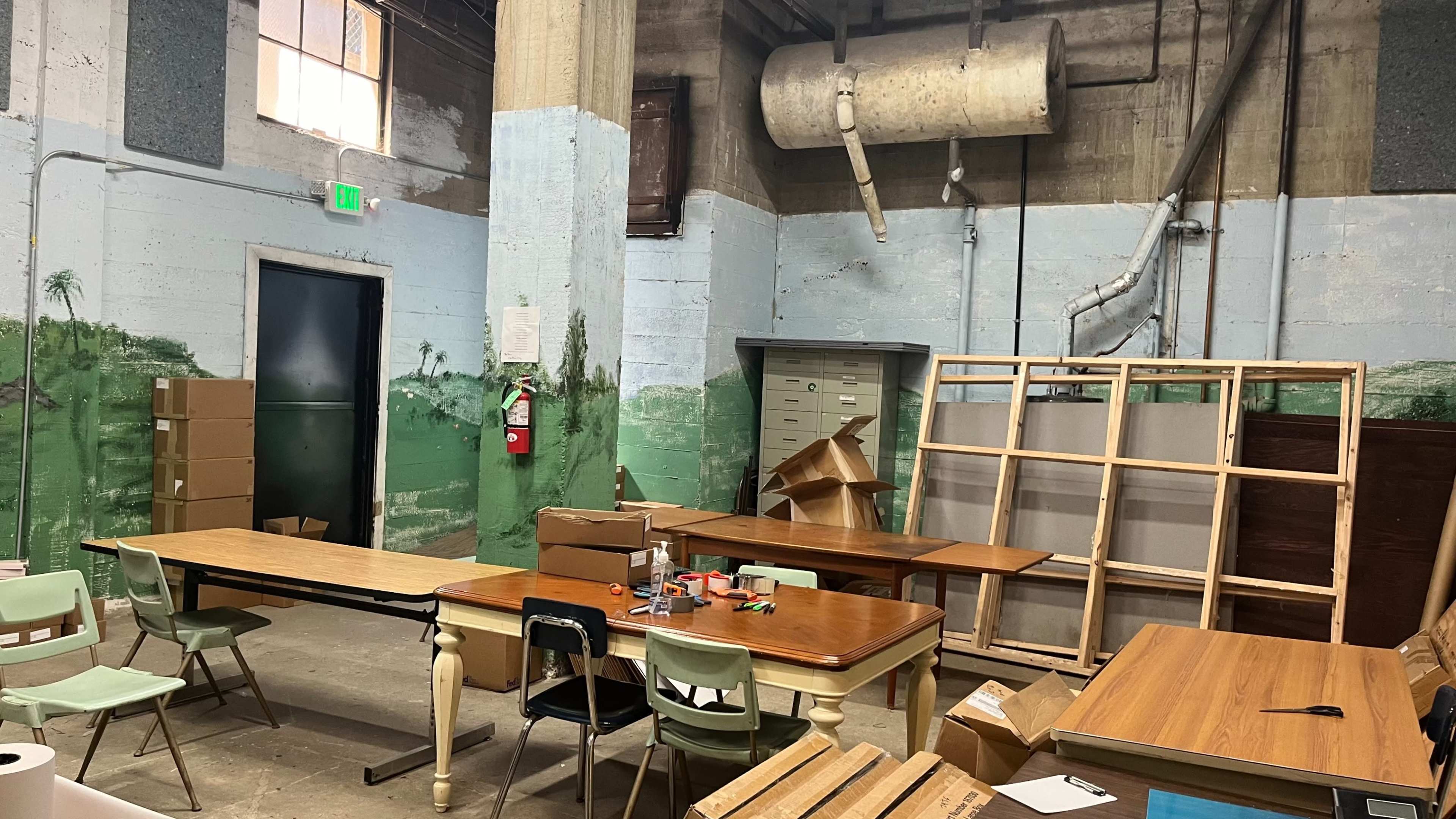 The image depicts a warehouse interior with several tables arranged in the center, cardboard boxes stacked nearby, and a wall featuring a painted mural of palm trees.