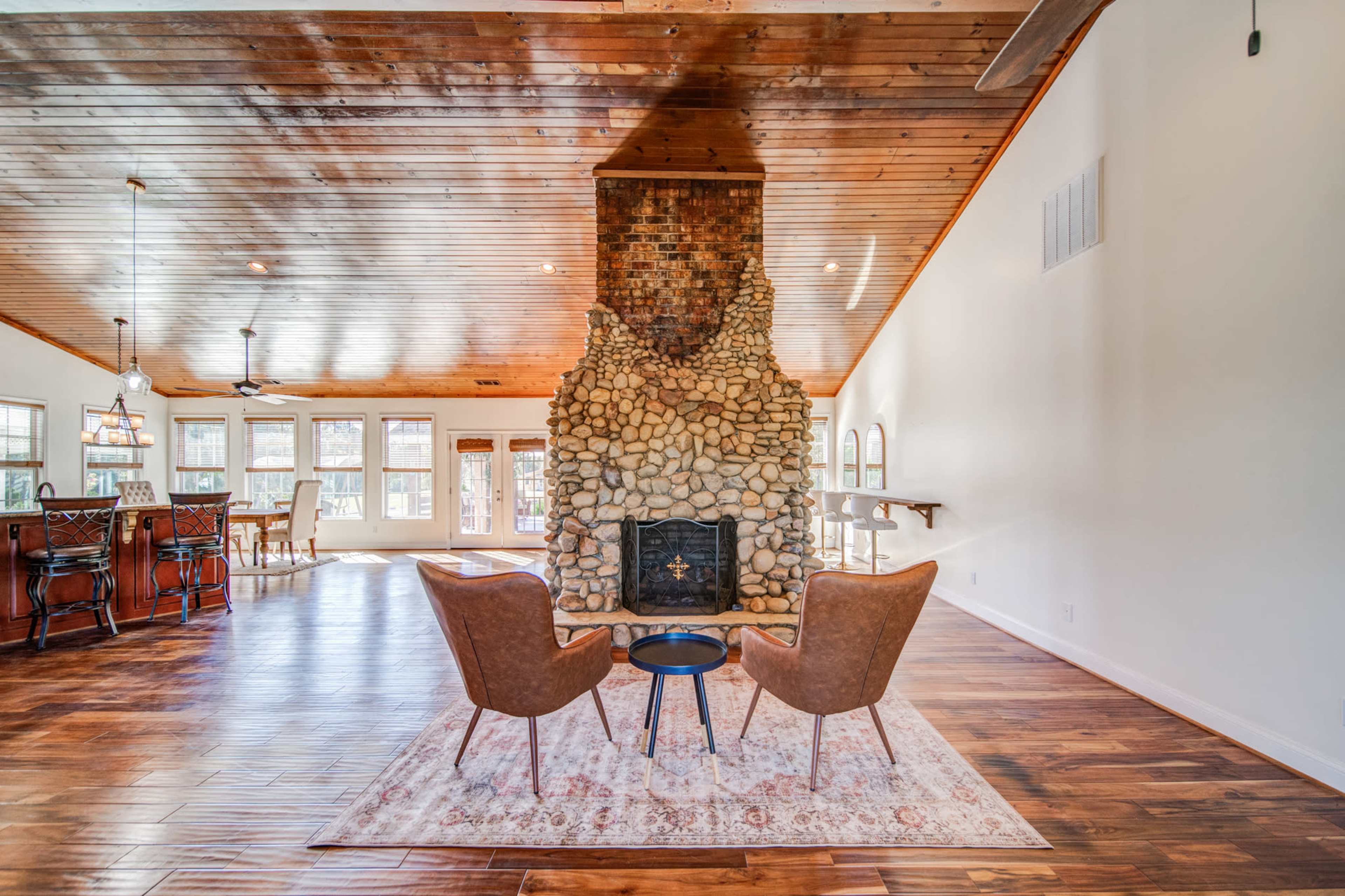 A spacious living room features a central stone fireplace surrounded by two leather chairs and a decorative rug.