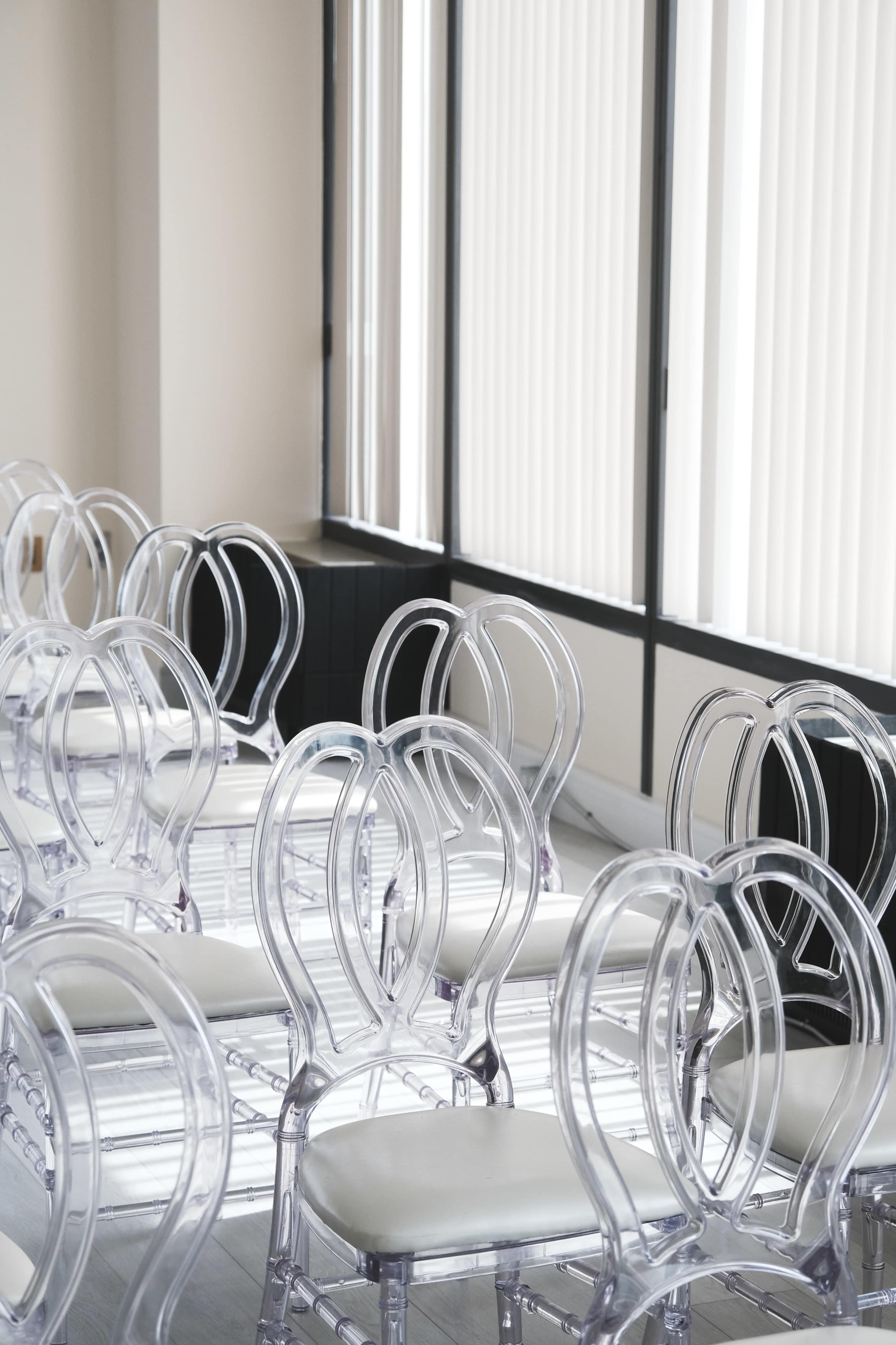 A row of transparent chairs with white cushions are set up in a well-lit room beside large windows with vertical blinds.