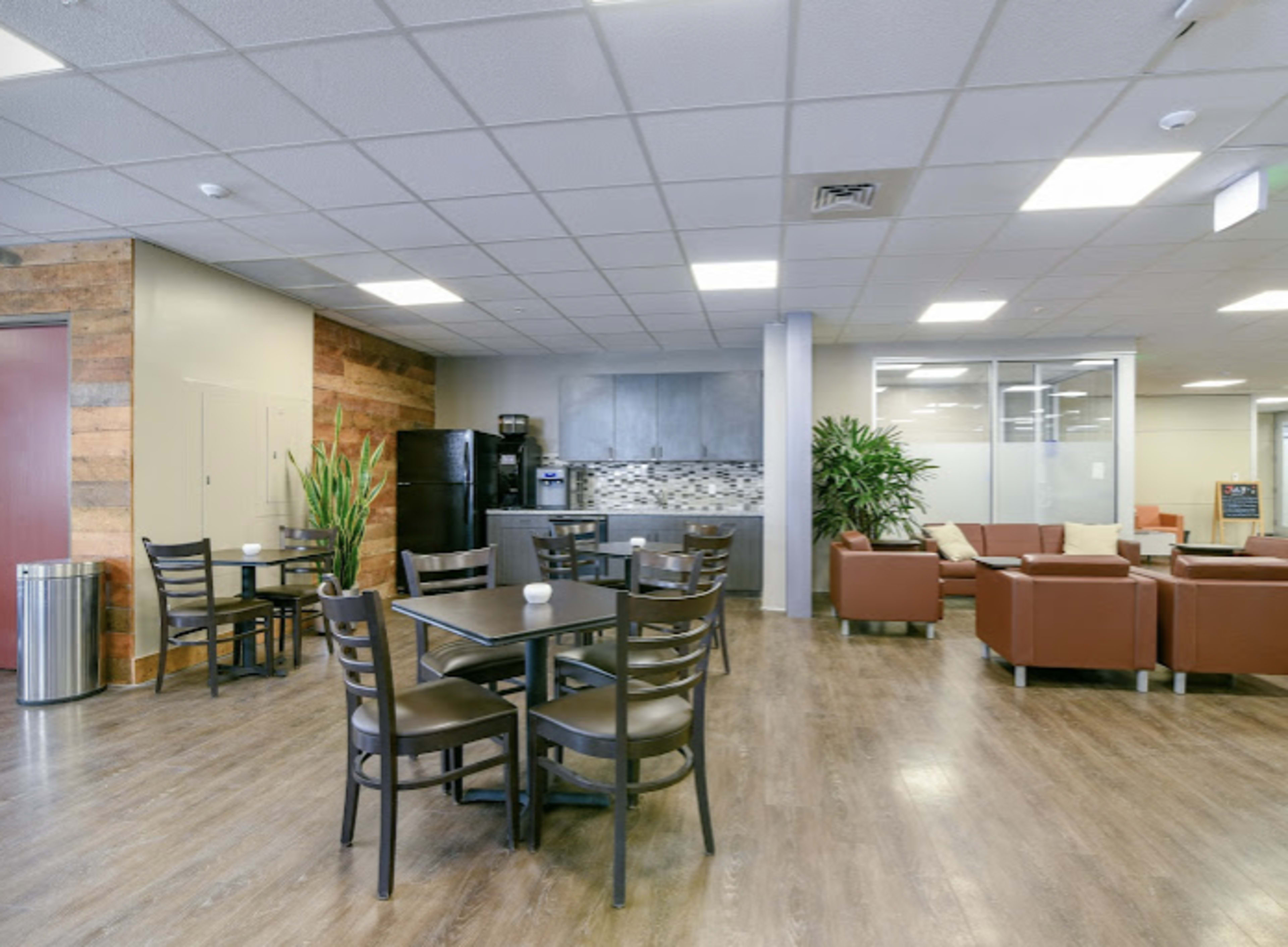 A modern lounge area features tables and chairs, a kitchen space with appliances, and comfortable seating arranged along a wall of plants.