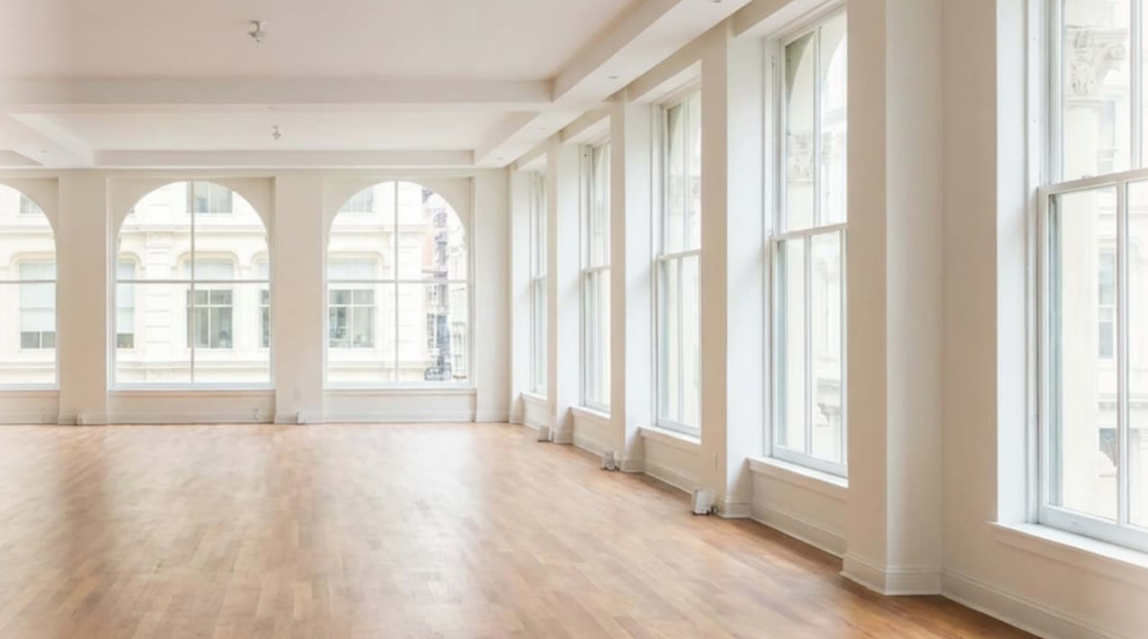 The image shows a spacious, well-lit room featuring large windows and a wooden floor.
