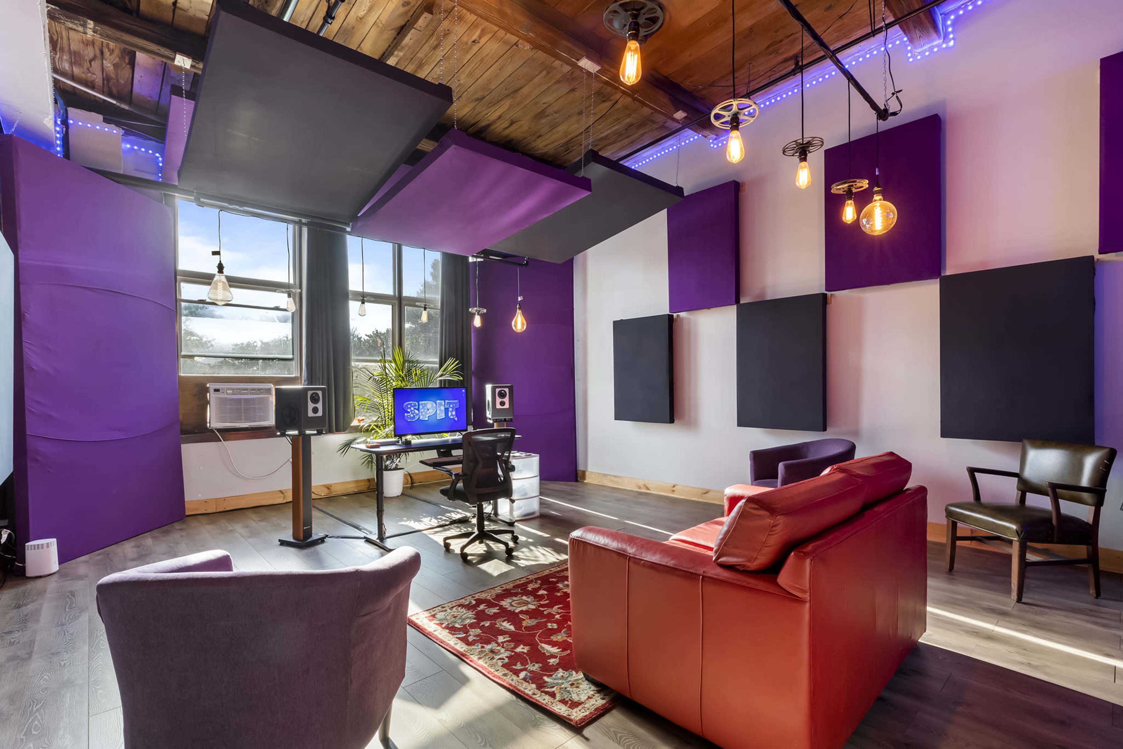 A modern recording studio features a red leather sofa, a desk with a computer, and soundproofing panels on the walls, with large windows allowing natural light to enter.