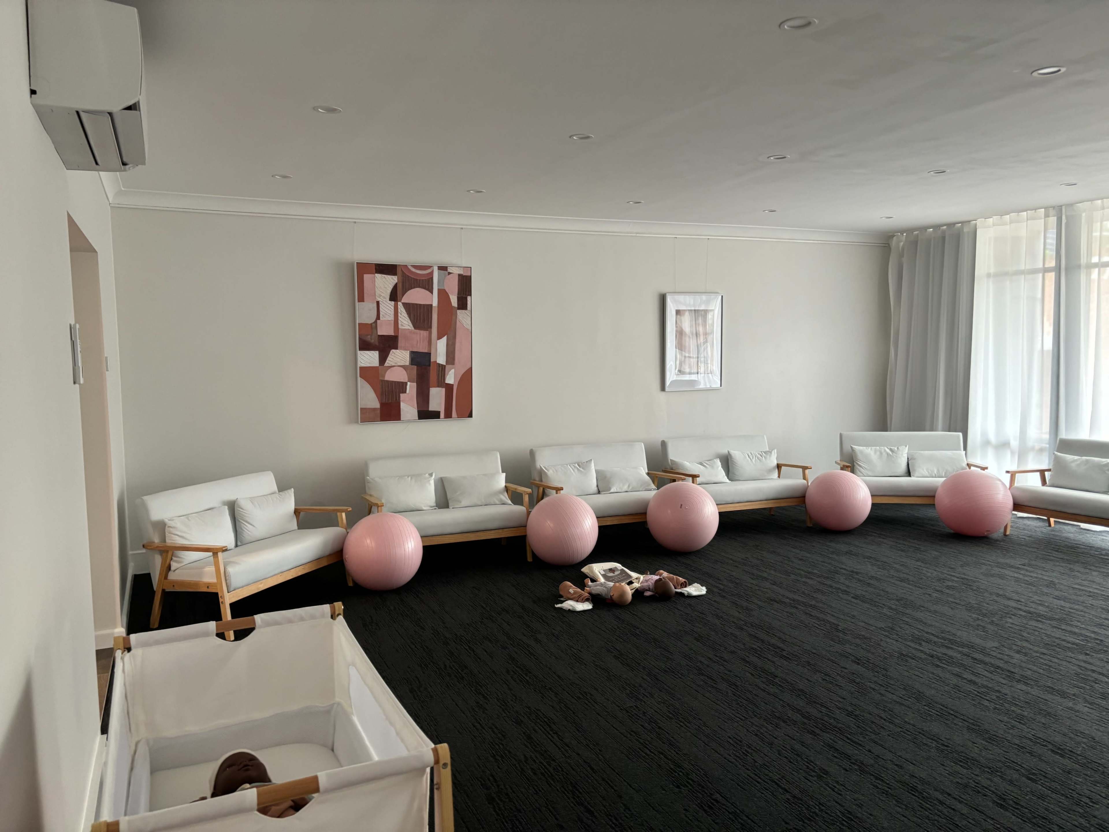The image shows a bright, minimalist room with several white chairs, pink exercise balls, and a crib in the corner.