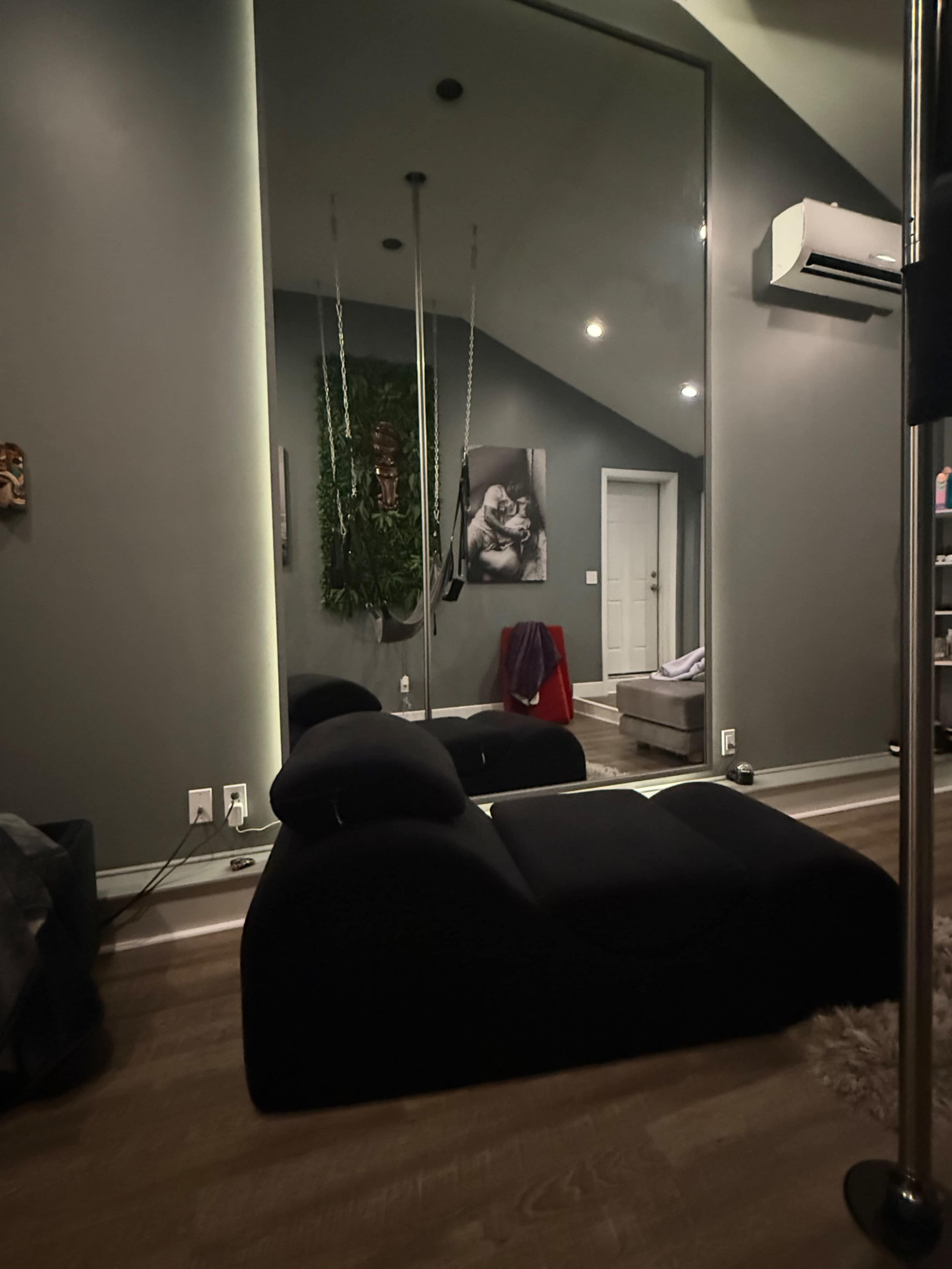 The image shows a modern room featuring a large mirror, a black lounge chair, and a wall adorned with greenery and contemporary artwork.