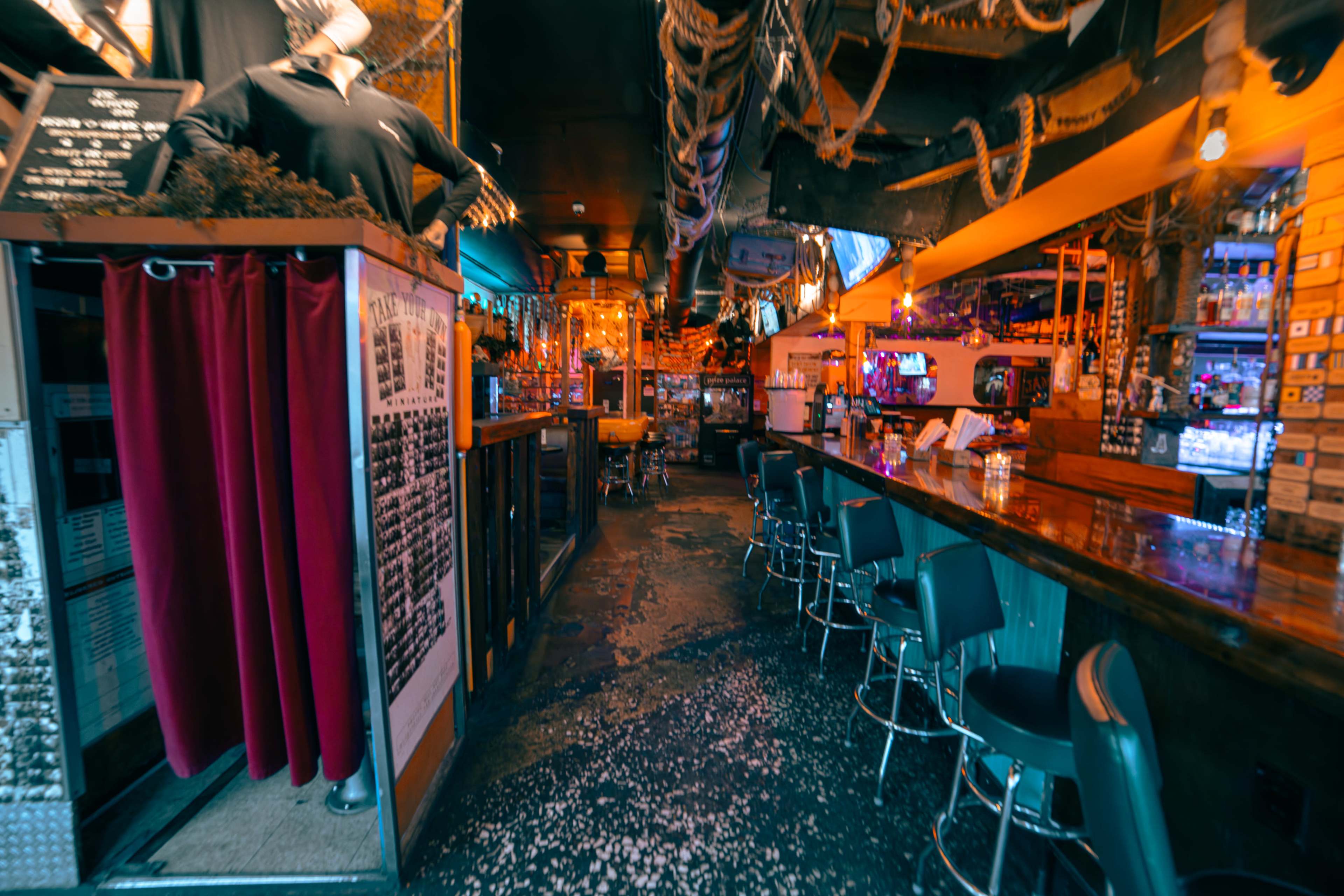 The image depicts the interior of a restaurant or bar with a long wooden counter, metal stools, and nautical-themed decor hanging from the ceiling.