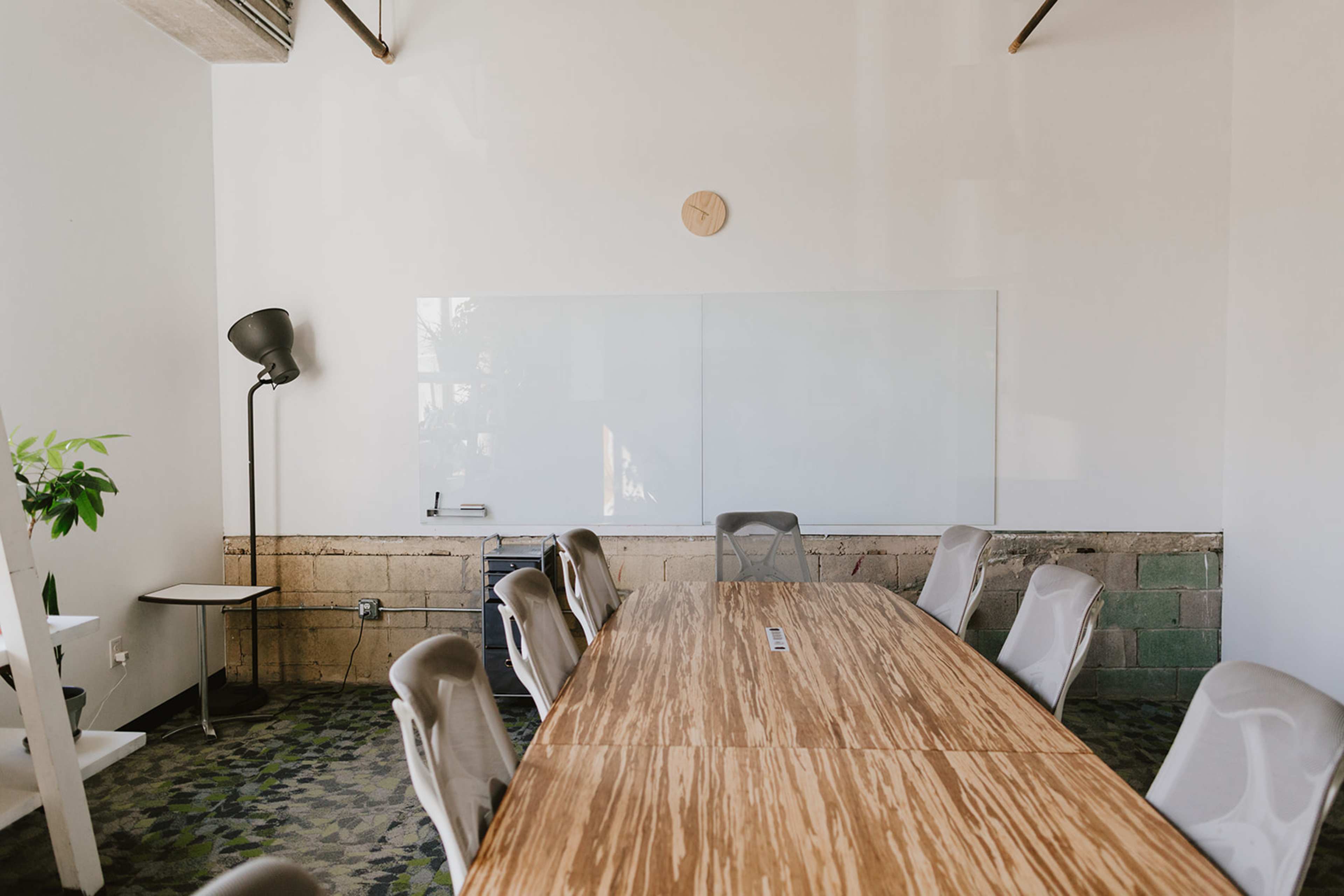 A modern conference room features a long wooden table surrounded by chairs, with a wall-mounted clock and a whiteboard in the background.
