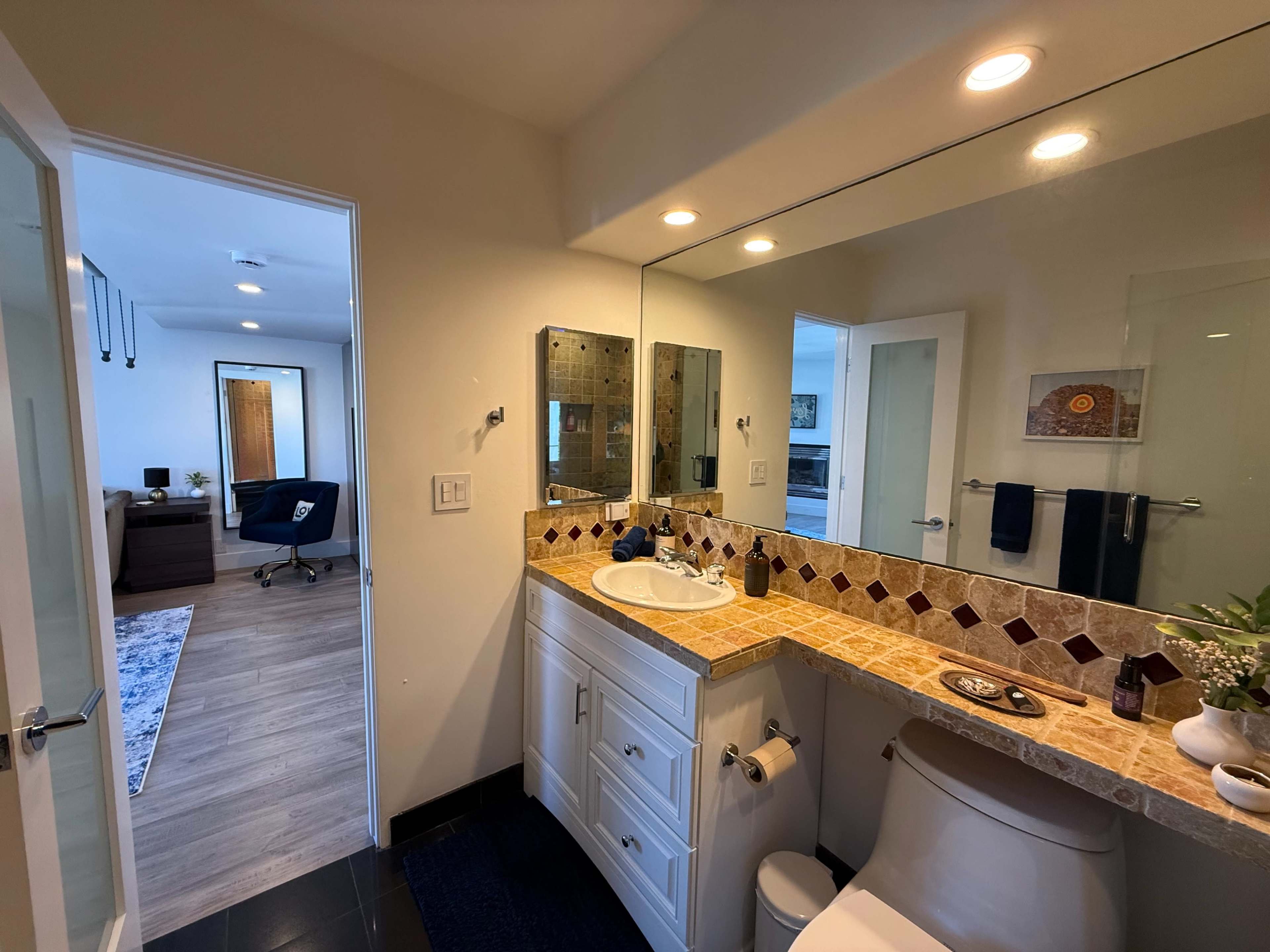 The image shows a bathroom with a tiled countertop, a mirror above the sink, and a view of an adjoining room through an open door.