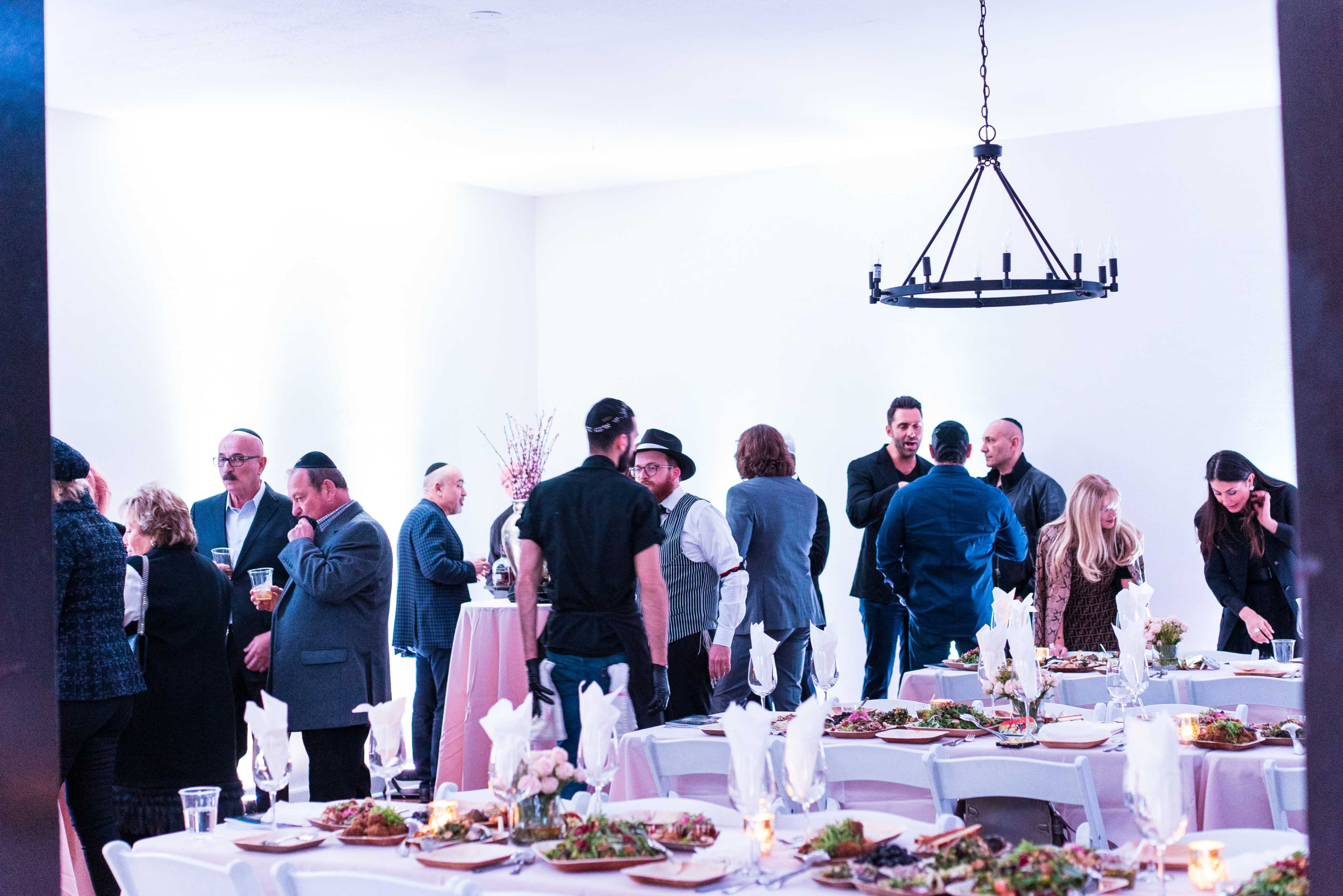 A group of people socializes in a well-lit space, with tables set for a meal and food displayed.