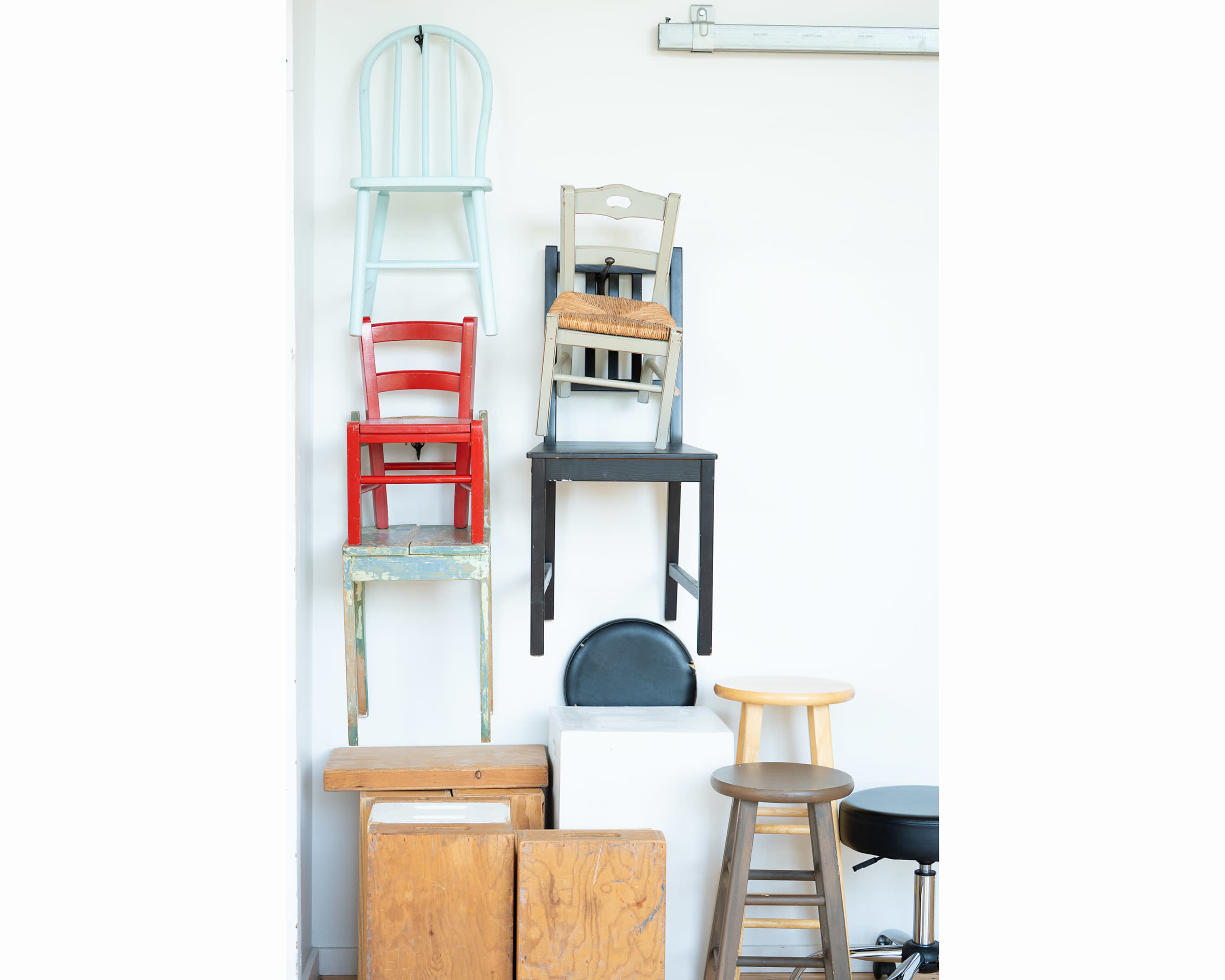 A variety of chairs, including wooden and metal types, are stacked against a white wall.