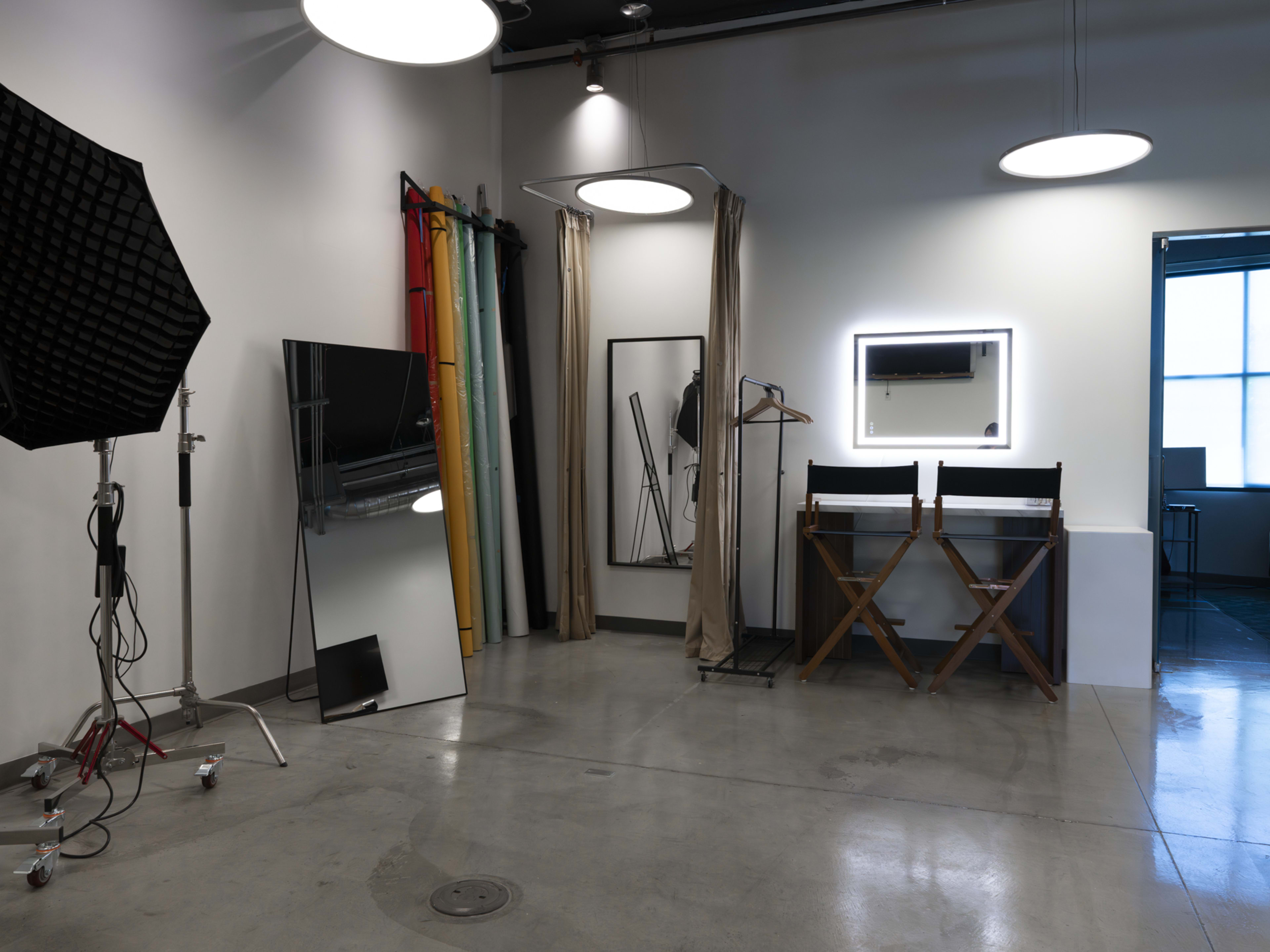 The image shows a well-equipped photography studio featuring a large mirror, a director's chair, a clothing rack, and backdrop rolls in various colors.