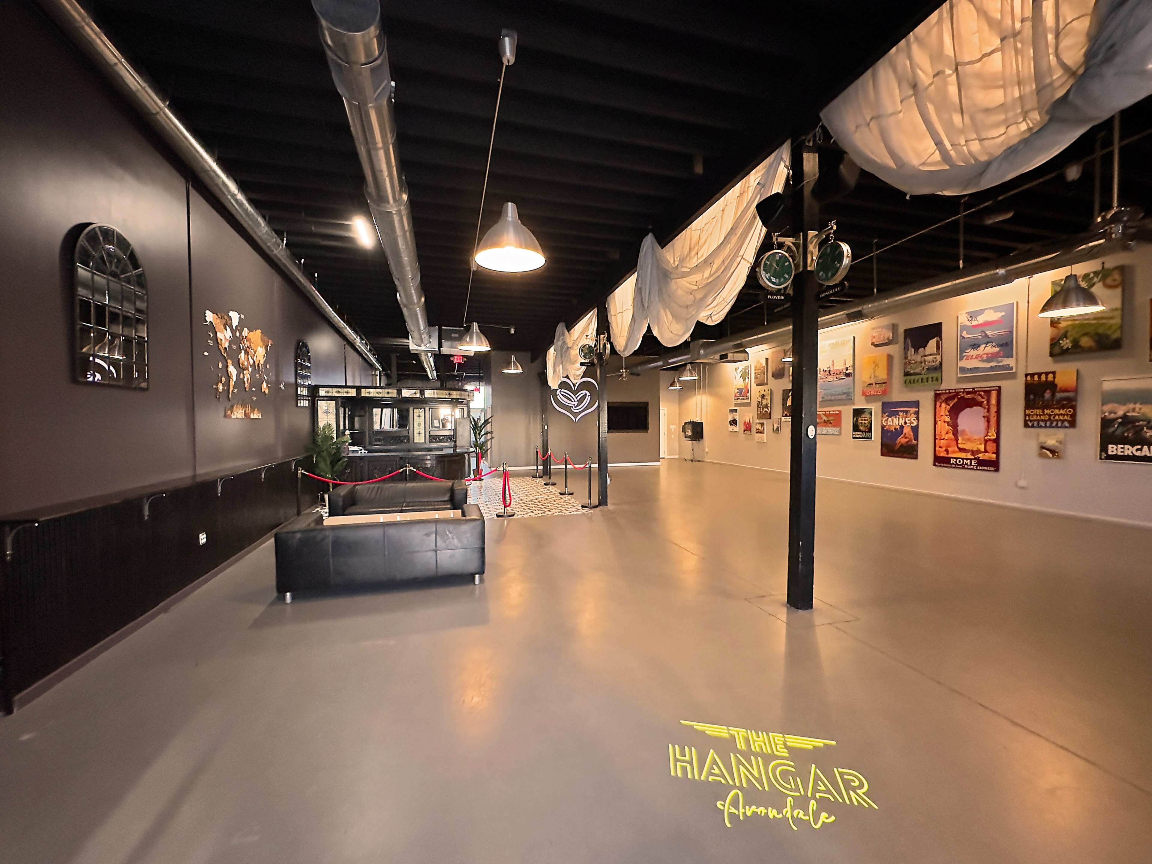 A spacious interior of an event venue called "The Hangar," featuring light-colored walls adorned with framed artwork and a central seating area with a black couch.