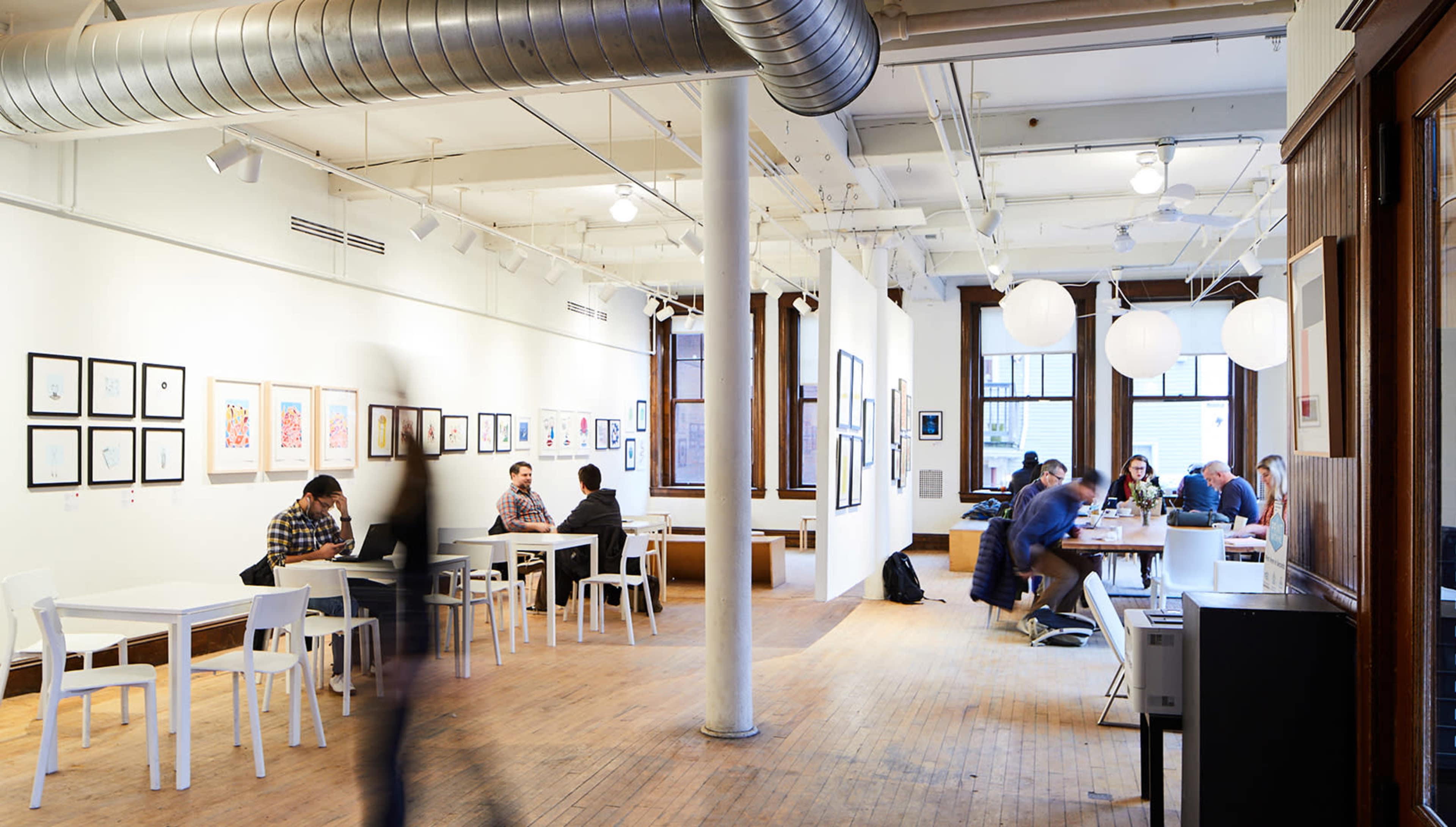 An art studio featuring tables occupied by people, with artwork displayed on the walls and large paper lanterns hanging from the ceiling.