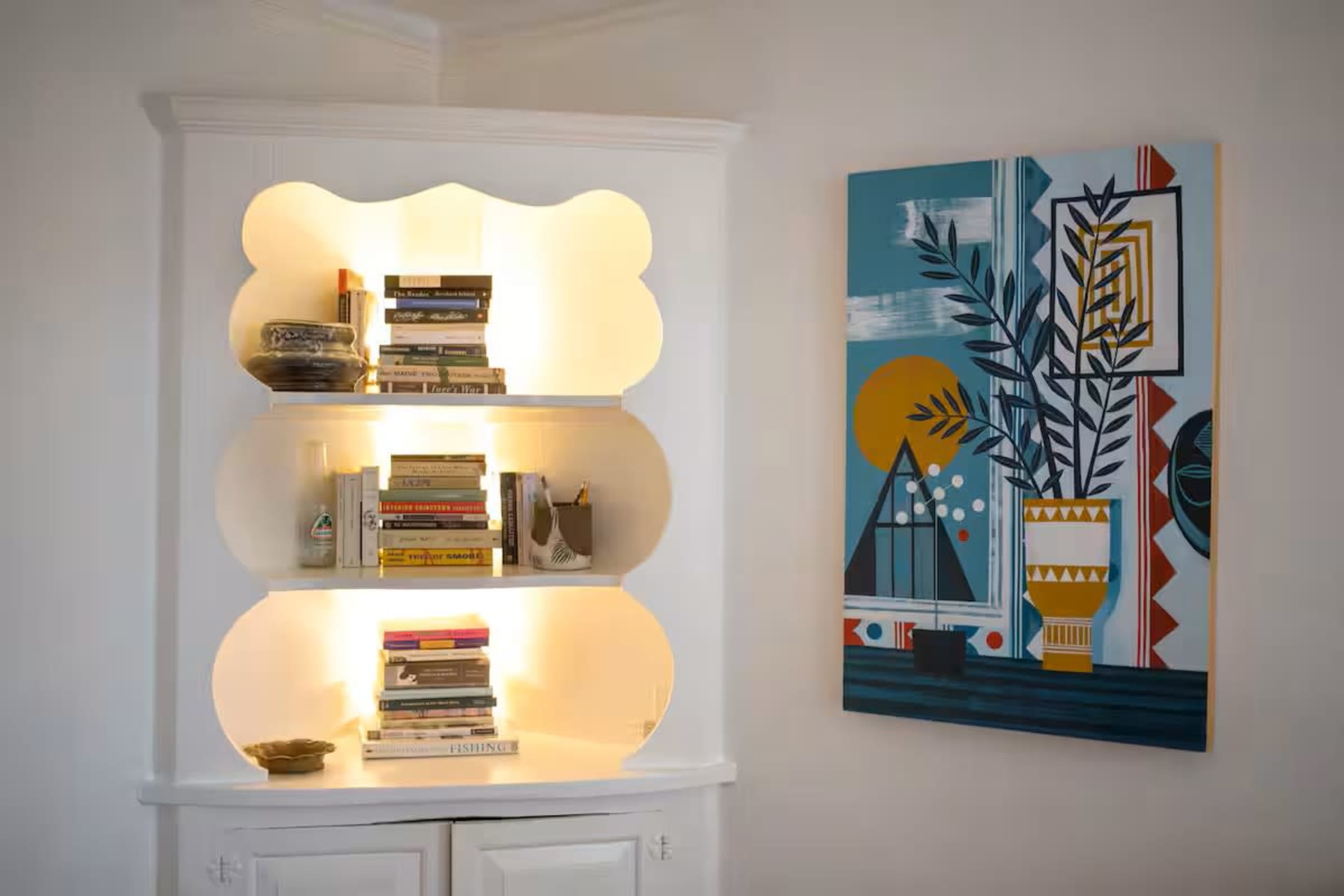 A white bookshelf with illuminated shelves holds various books, and a colorful abstract painting featuring geometric shapes is hung on the wall beside it.