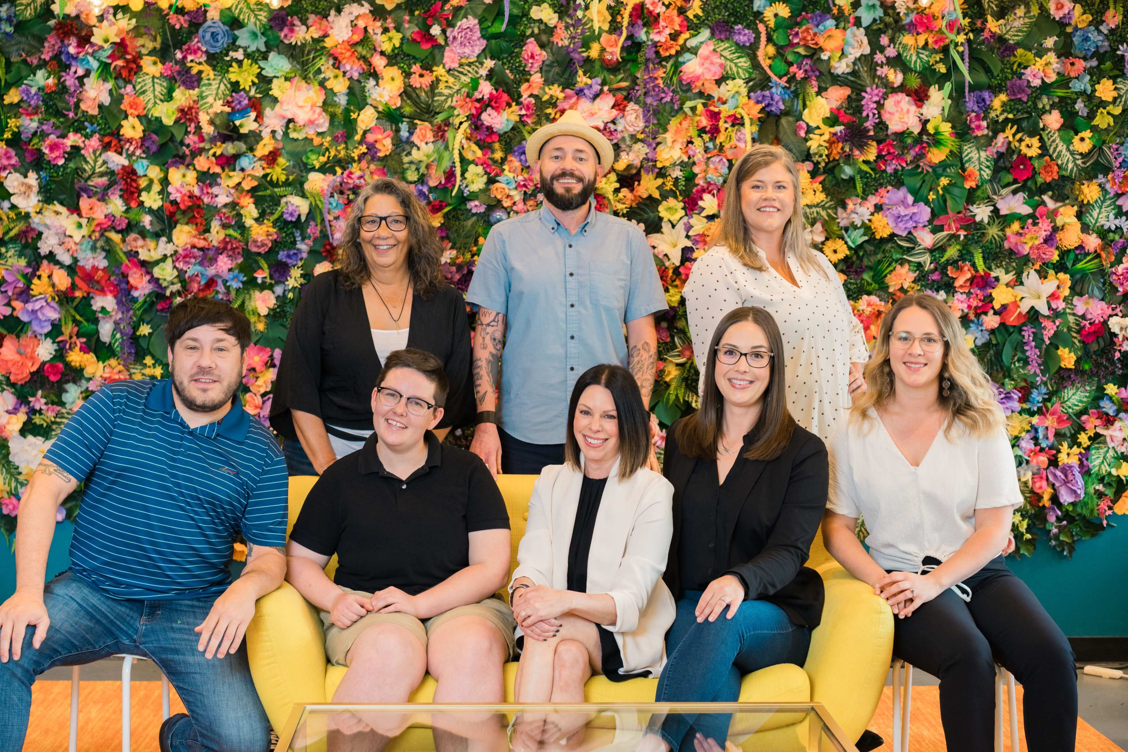 A group of eight individuals poses together in front of a vibrant floral wall, seated on a yellow sofa.