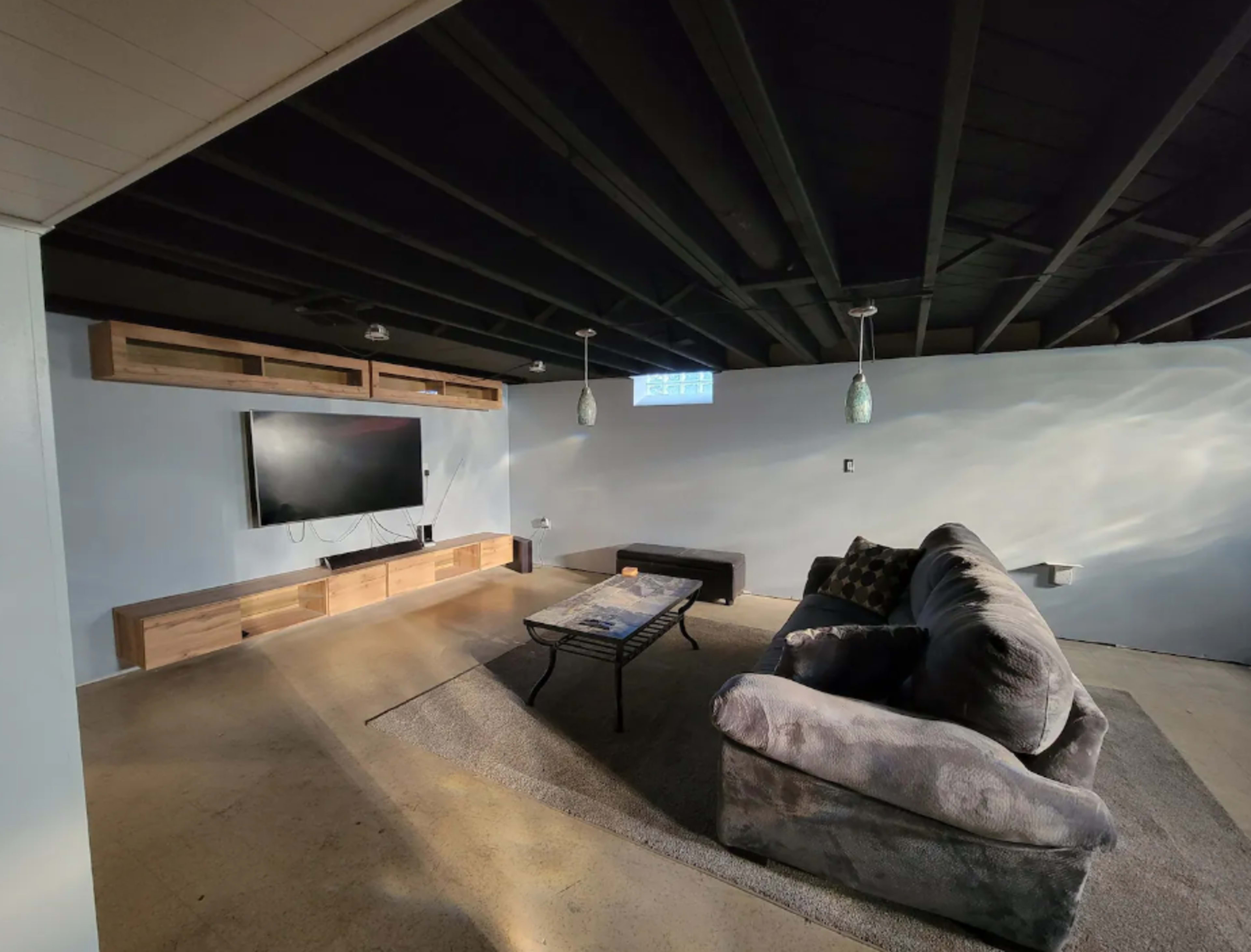 The image shows a basement living area with a large sectional couch, a coffee table, a mounted television, and wooden shelving against the wall.