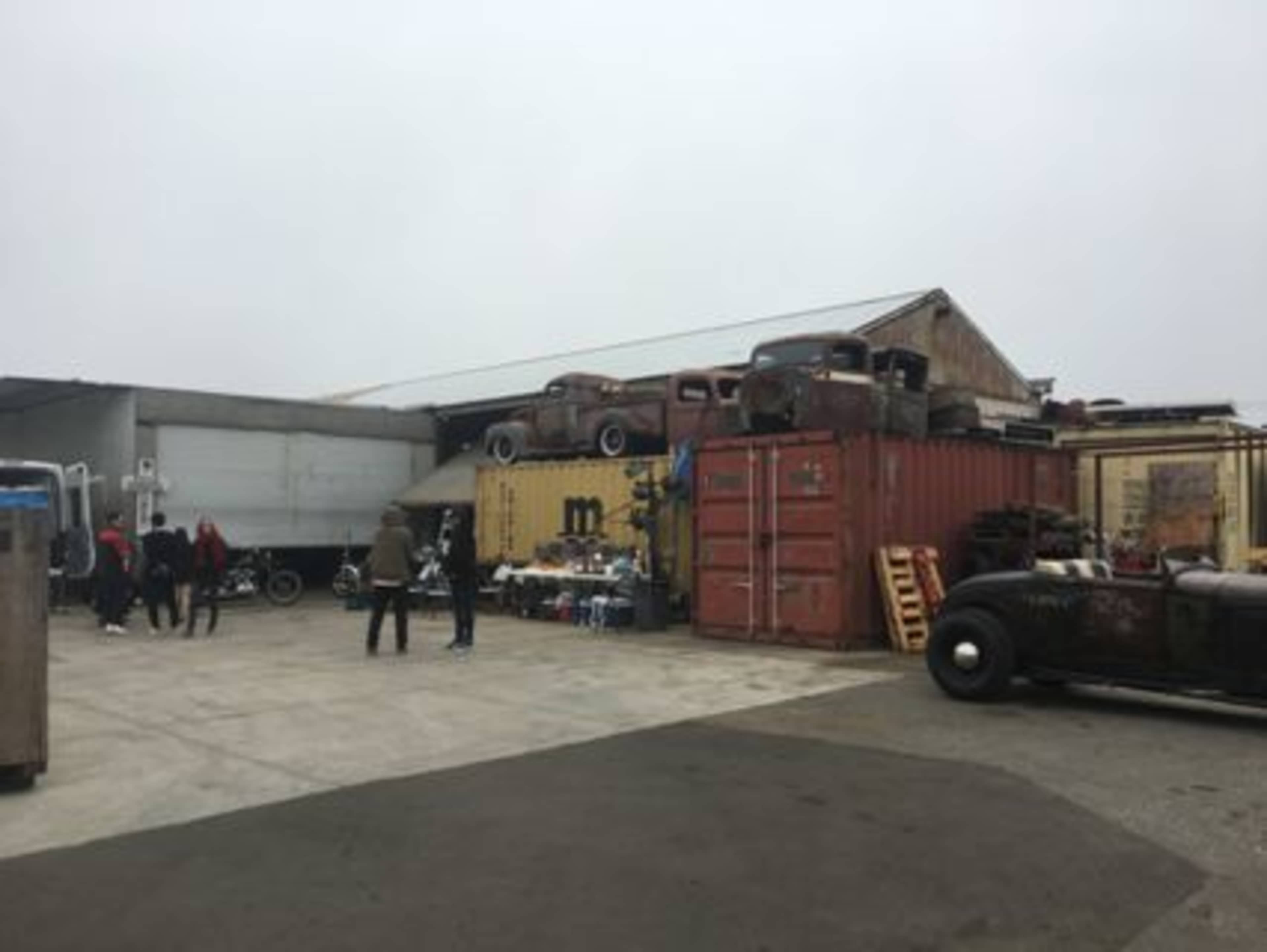 A collection of vintage cars and shipping containers is displayed in an industrial yard under a cloudy sky.