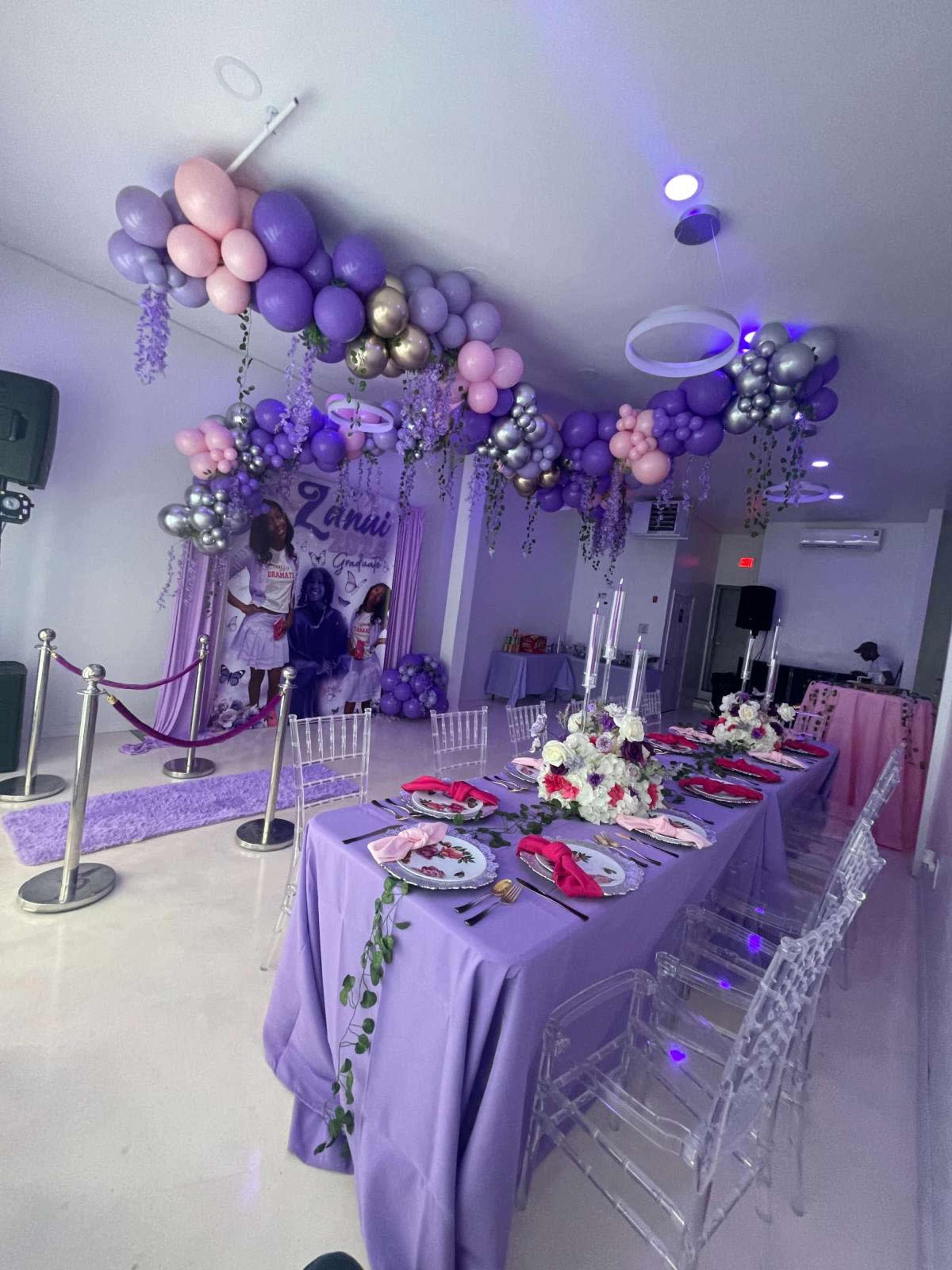 The image depicts a festive indoor event space decorated with purple and pink balloons, a long dining table set with vibrant tableware, and elegant floral arrangements.