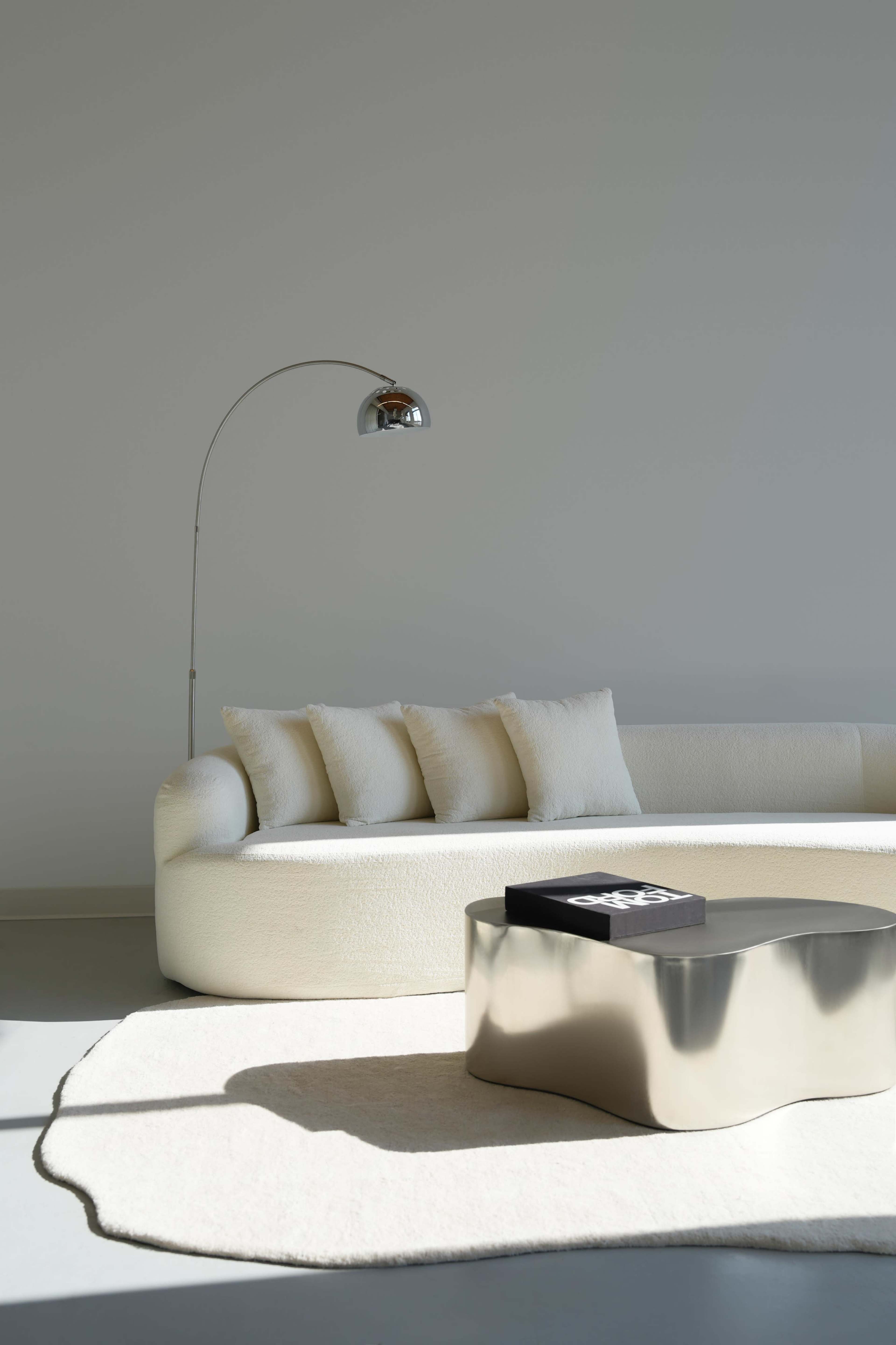 A curved white sofa with three pillows is positioned next to a metallic coffee table, while an arc floor lamp stands nearby on a light-colored rug.