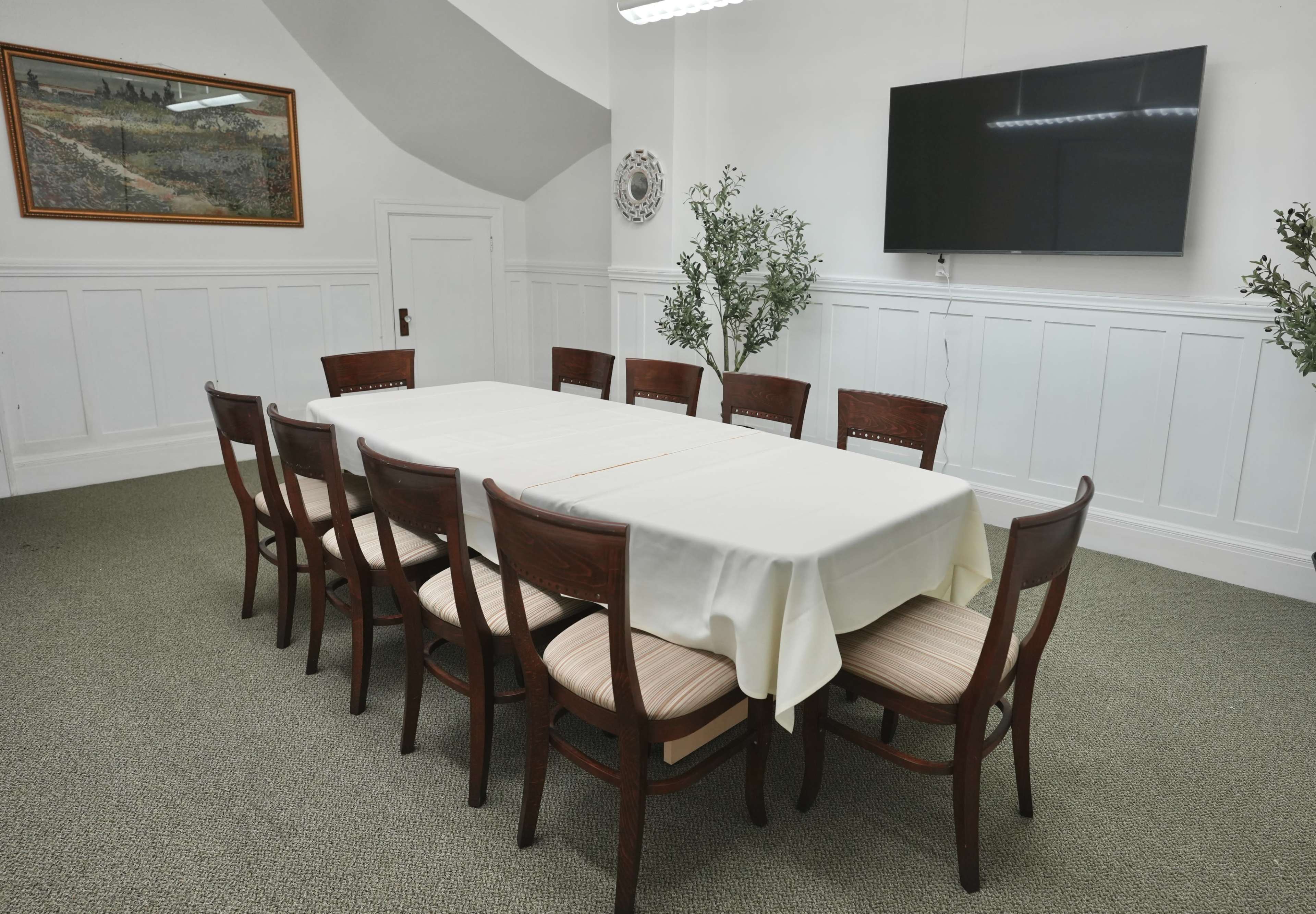A long table covered with a white tablecloth is surrounded by twelve wooden chairs in a well-lit meeting room featuring a wall-mounted screen and a painting.