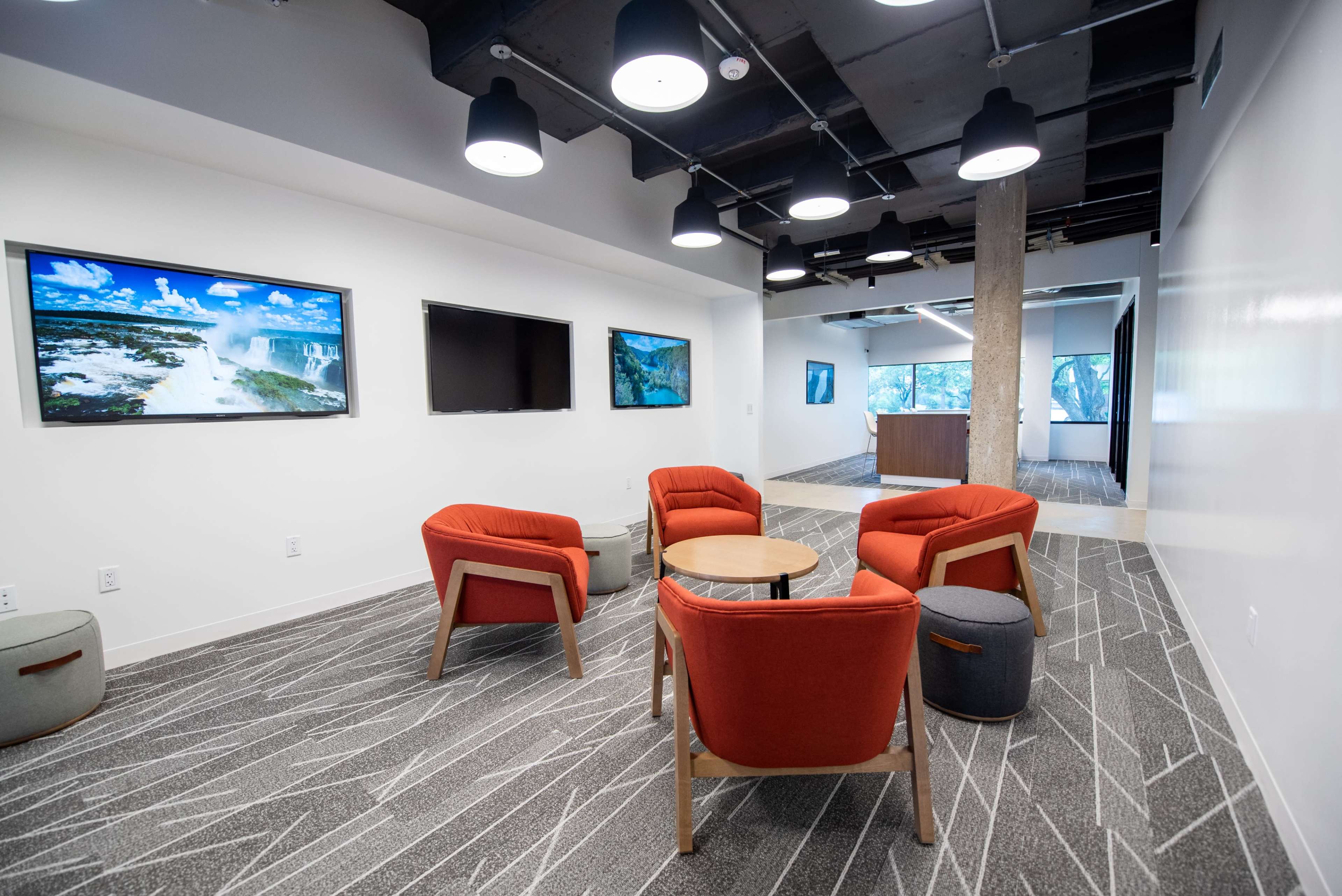 A modern office space with four red chairs arranged around a wooden table, two wall-mounted screens displaying images, and a reception area visible in the background.