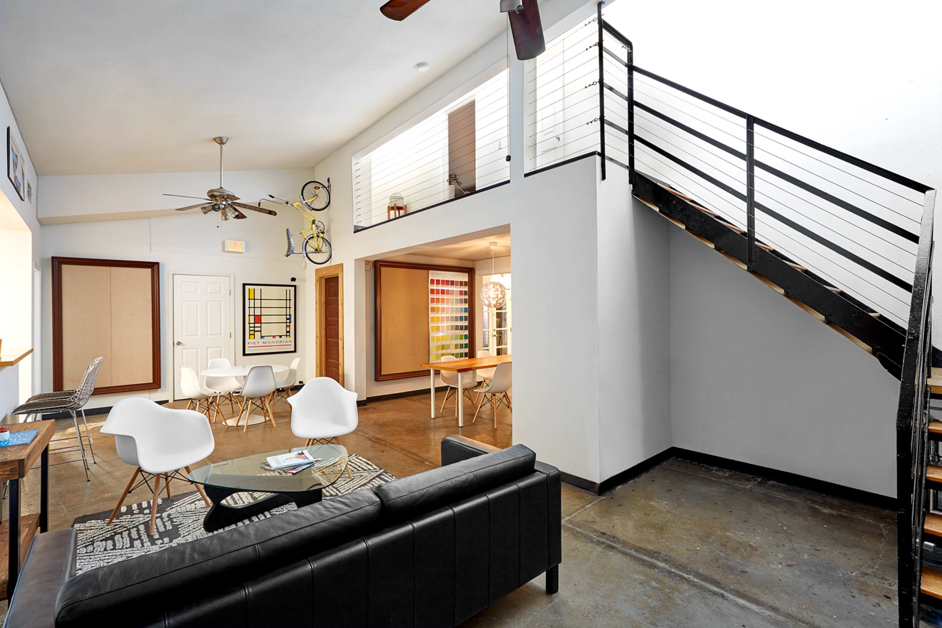A modern living space with a black sofa, a glass coffee table, and a staircase leading to an upper level, featuring an open layout and a view of a colorful wall.