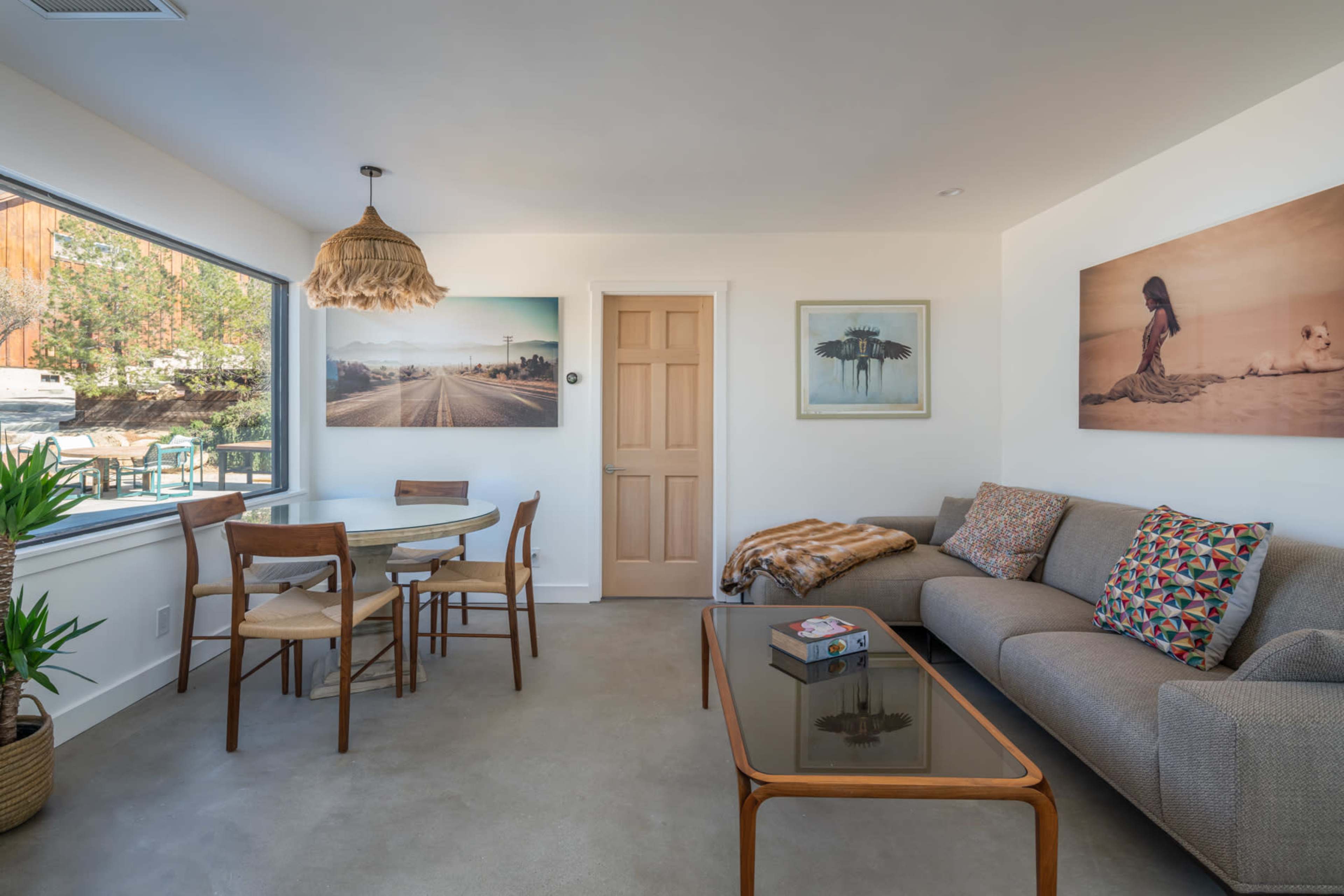 A modern living room with a sofa, a glass coffee table, a dining table with chairs, and artwork on the walls, all illuminated by natural light from a large window.