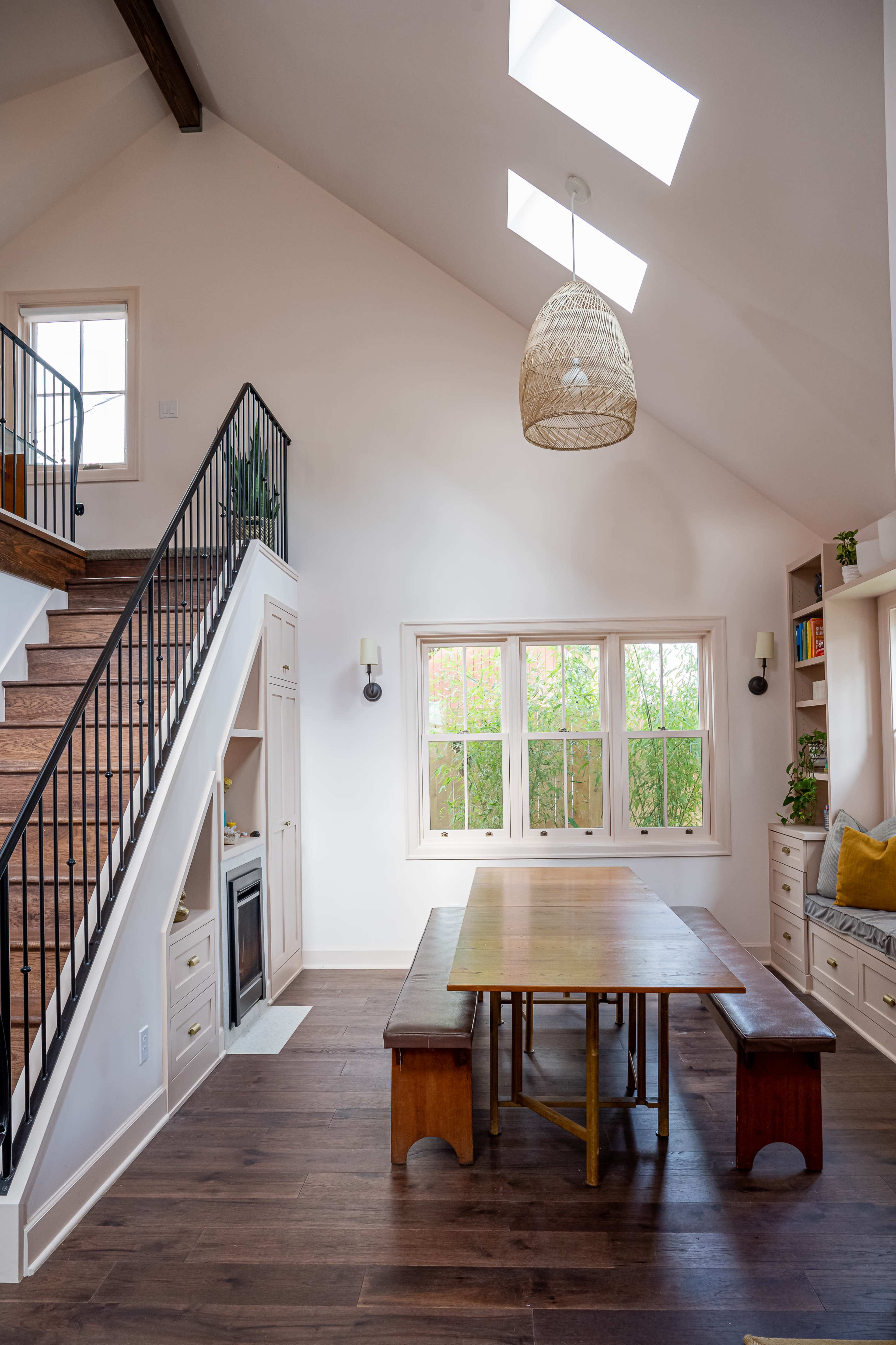 A dining area with a large wooden table and benches is located beneath a sloped ceiling, with a staircase nearby and large windows allowing natural light to enter.
