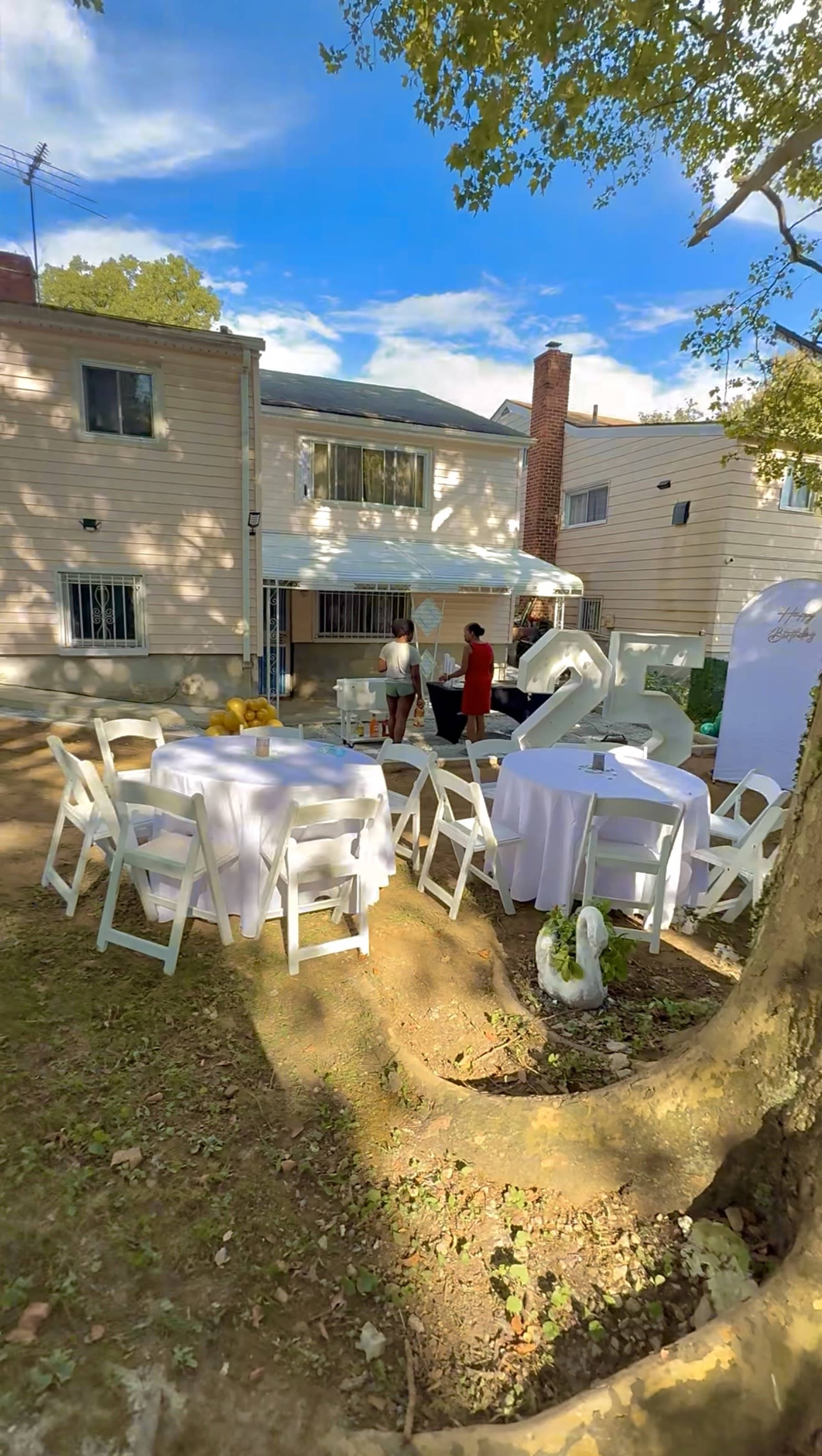 The image shows a backyard setting with white tables and chairs arranged for a gathering, while two people prepare decorations near a tent.