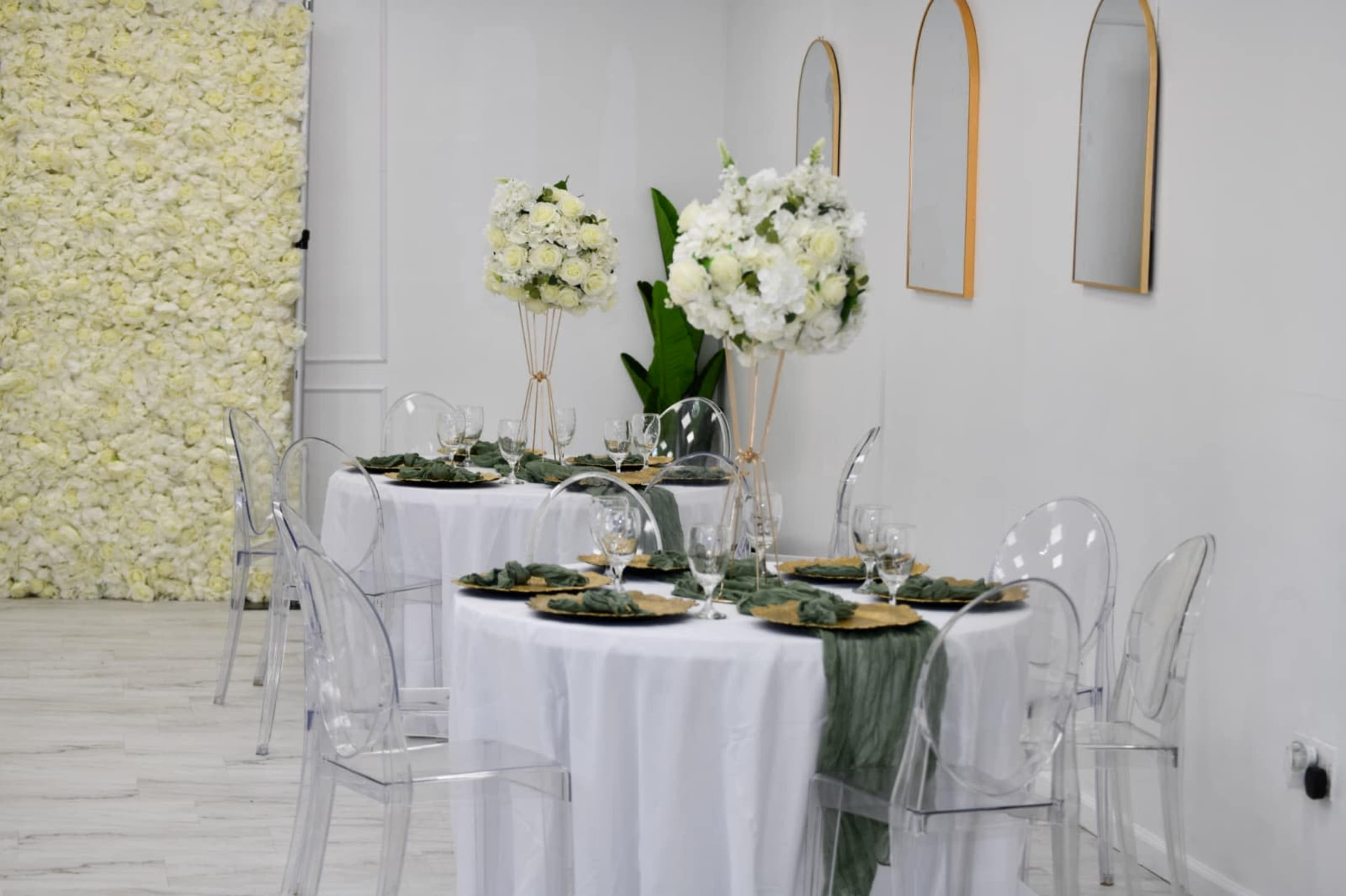 The image shows a beautifully arranged dining area with two round tables, each topped with elegant table settings, glassware, and floral centerpieces, against a backdrop of a flower wall and decorative mirrors.