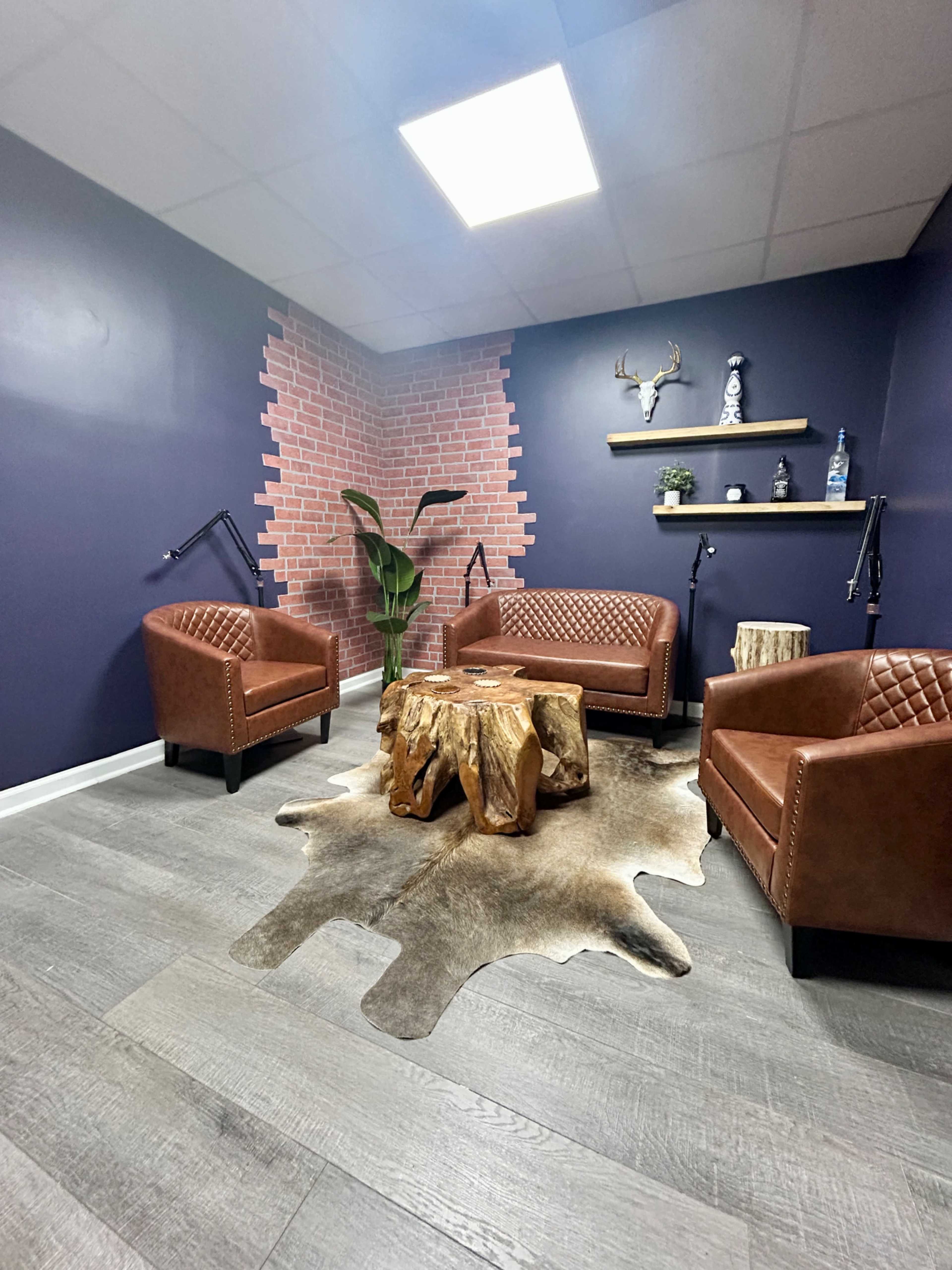 The image shows a small interior space with brown leather chairs, a wooden coffee table, a plant, and a wall featuring a brick design.
