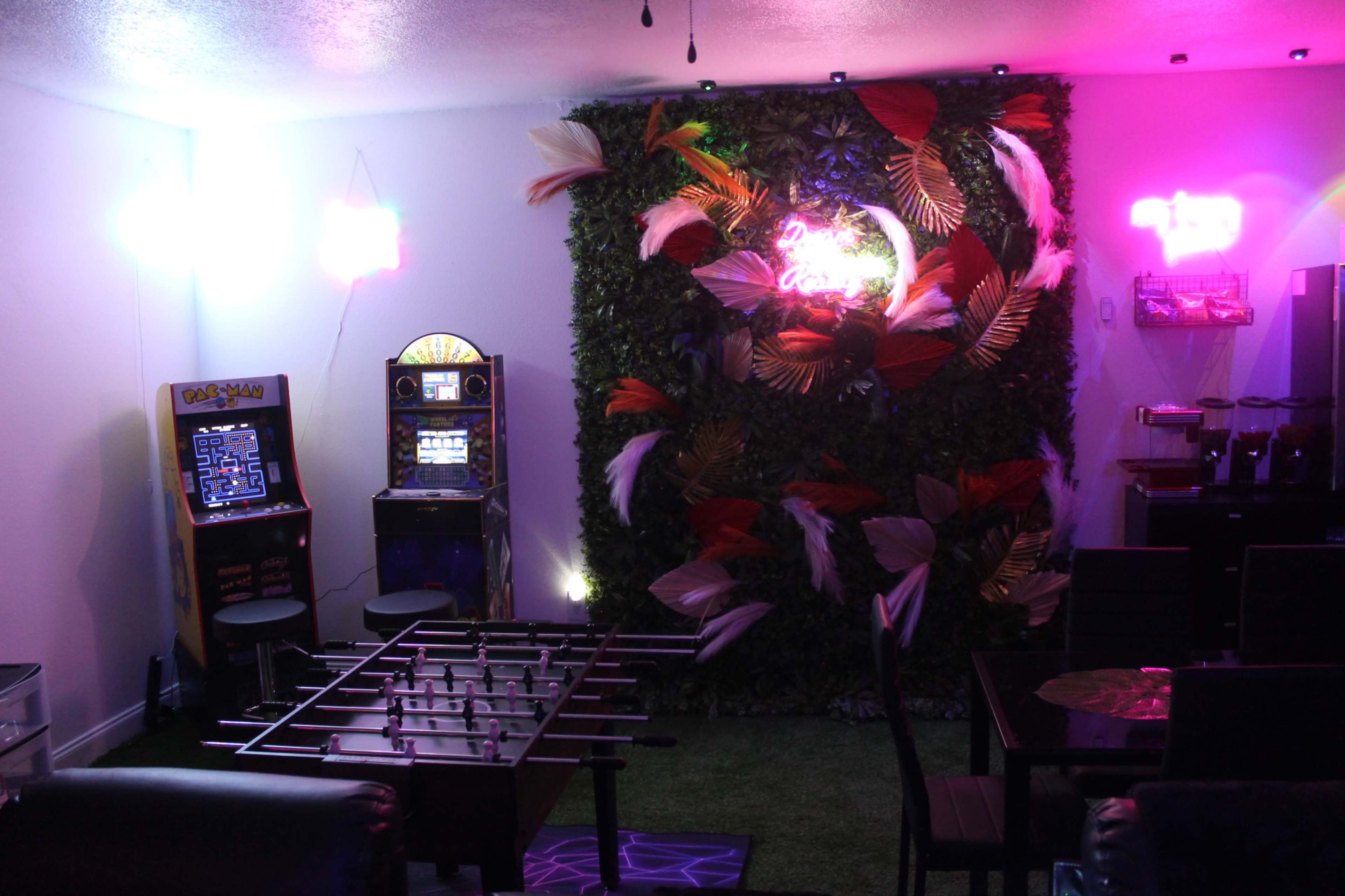 A game room with a foosball table, a classic arcade machine, and a vibrant wall adorned with artificial foliage and bright neon lights.