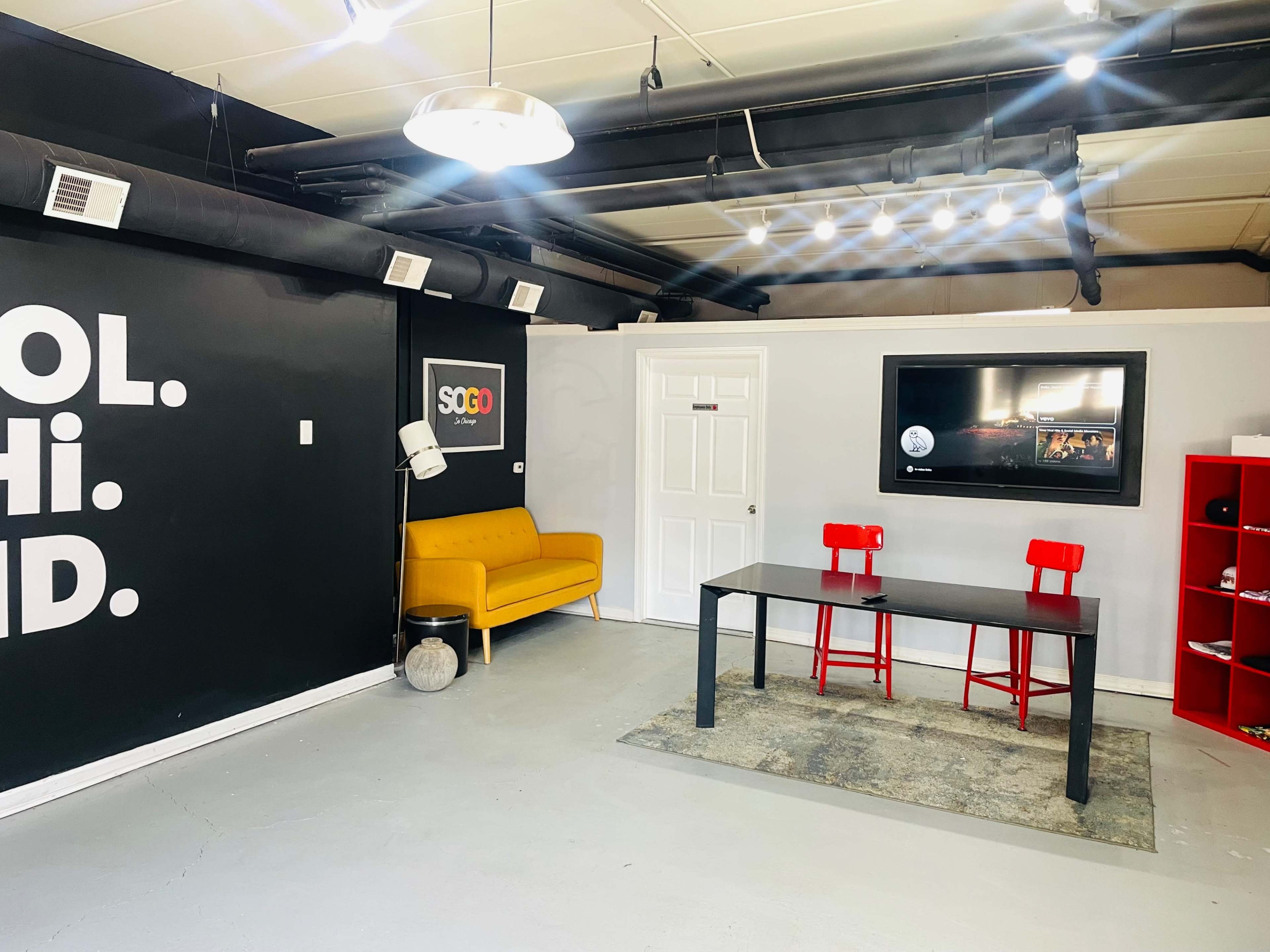 A modern indoor space featuring a yellow sofa, a black table with two red chairs, a wall with the words "SCHOOL HI. DID." in bold letters, and a television mounted on the wall.