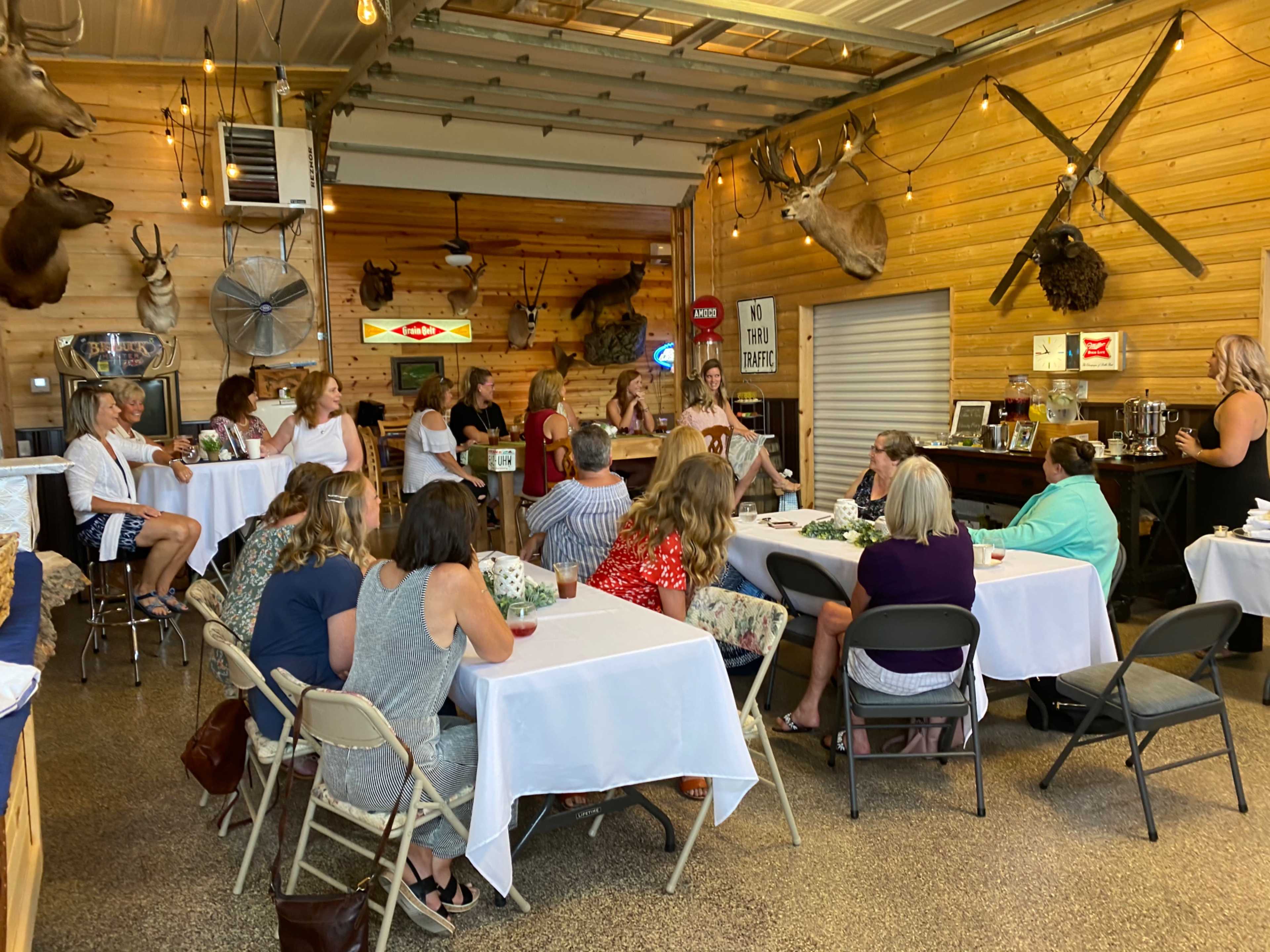A group of individuals sits around tables in a wooden-paneled room decorated with mounted trophies and vintage signs.