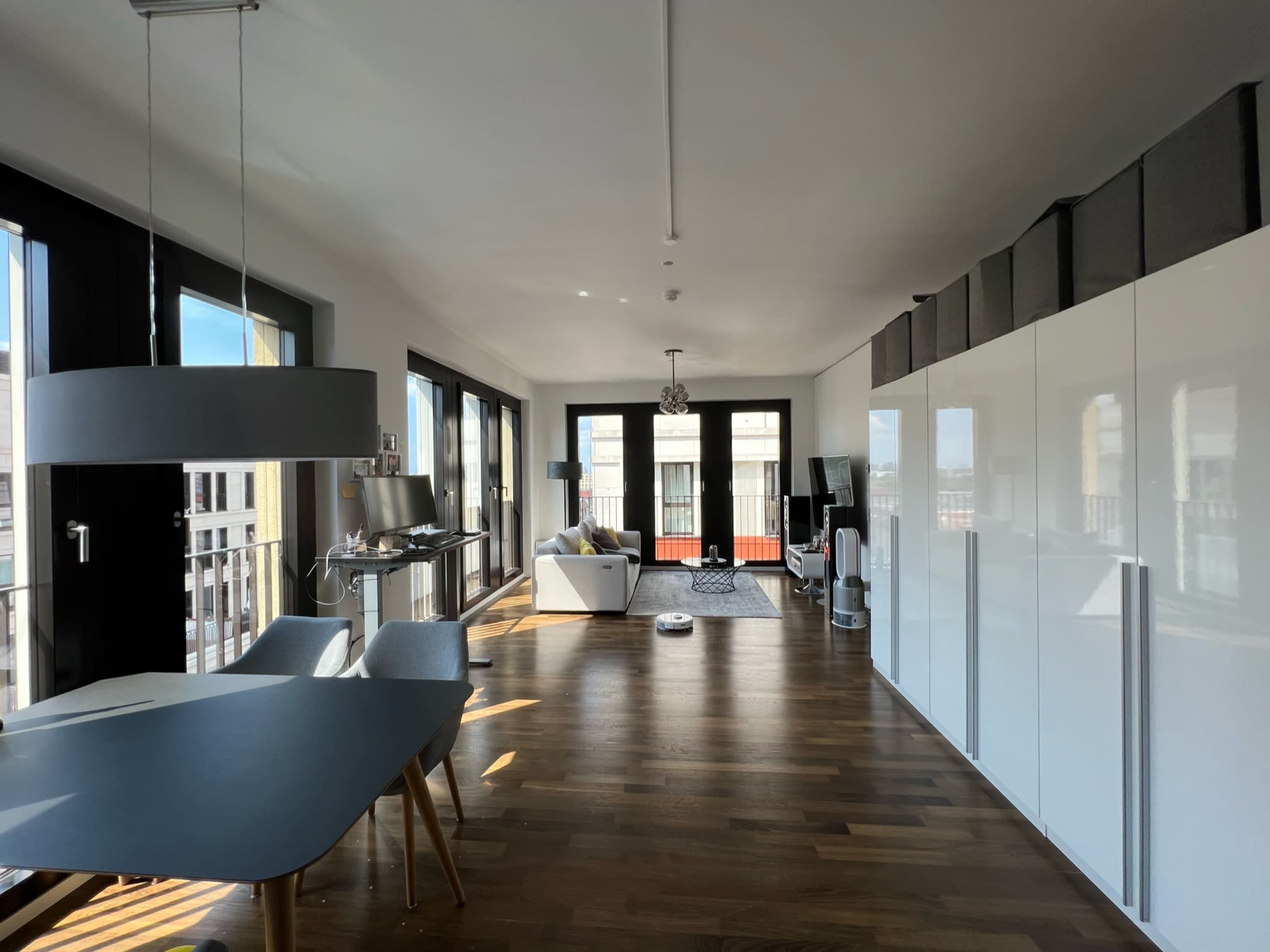 A modern, open living space with a dining area featuring a table and chairs, a white sofa, and large windows allowing natural light.