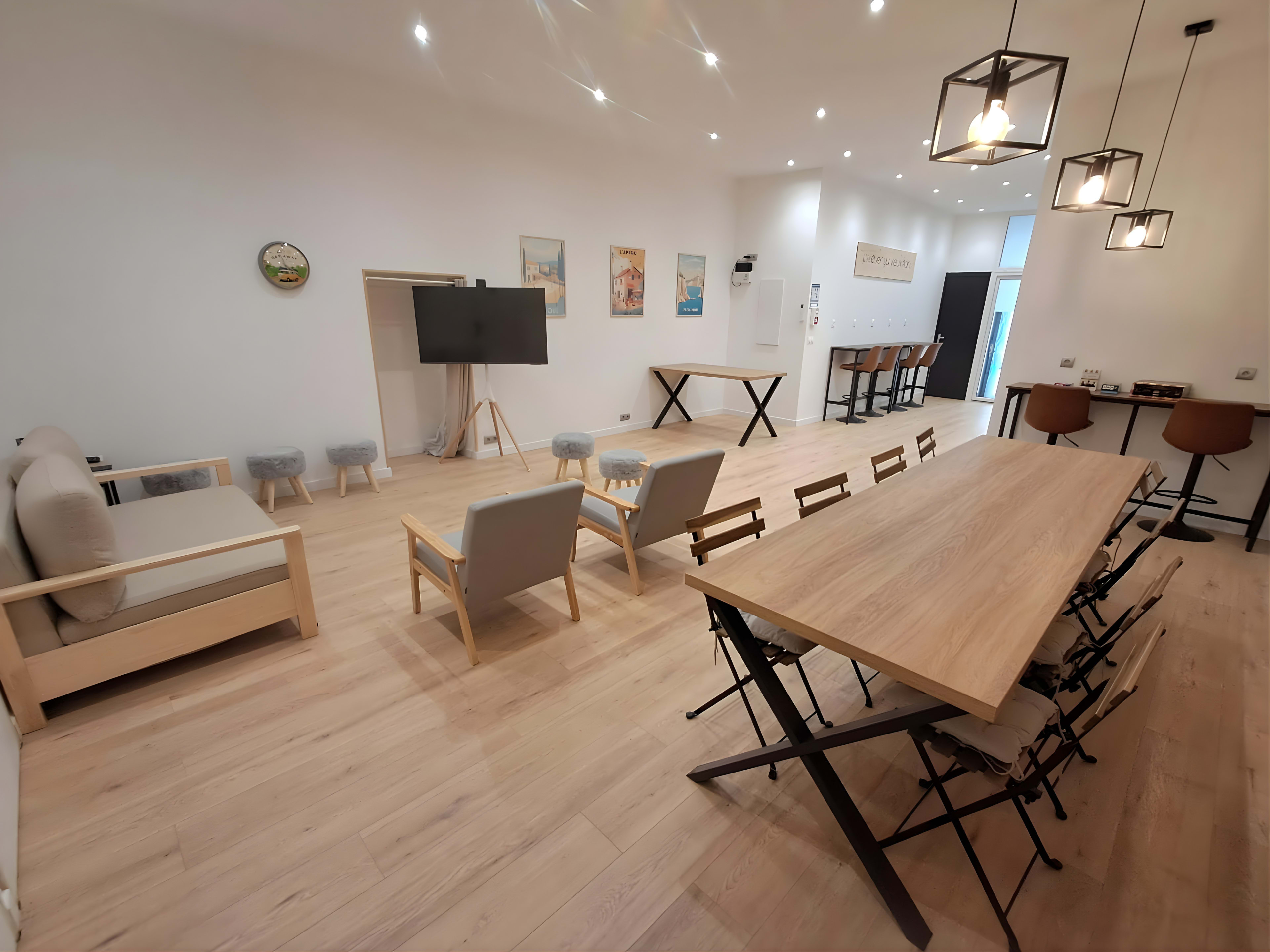 The image shows a modern, spacious room with light wooden floors, a large wooden table, seating areas, a television, and decorative artwork on the walls.