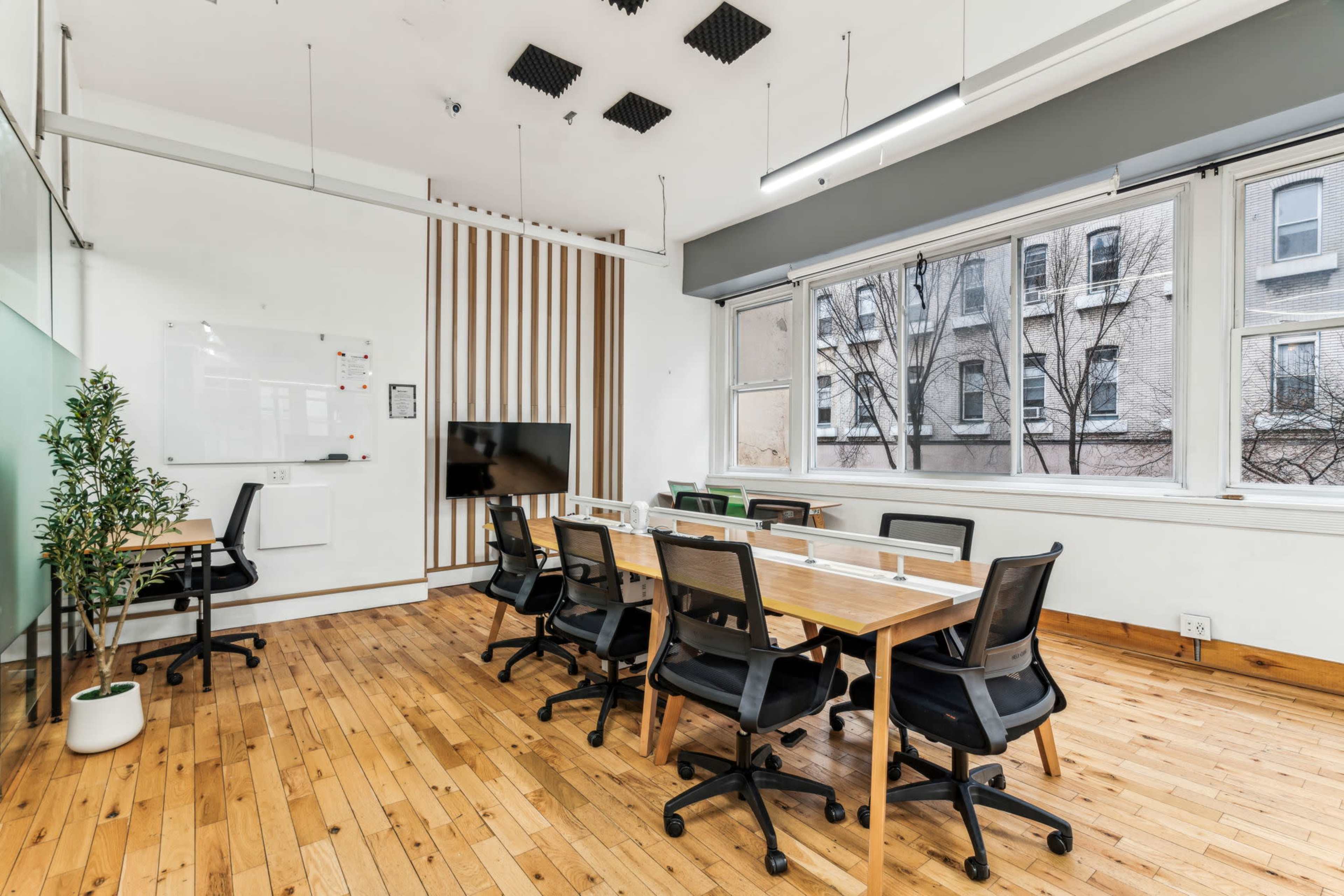 A modern conference room features a large table surrounded by black chairs, a whiteboard, and a wall-mounted screen, with wooden flooring and ample natural light.