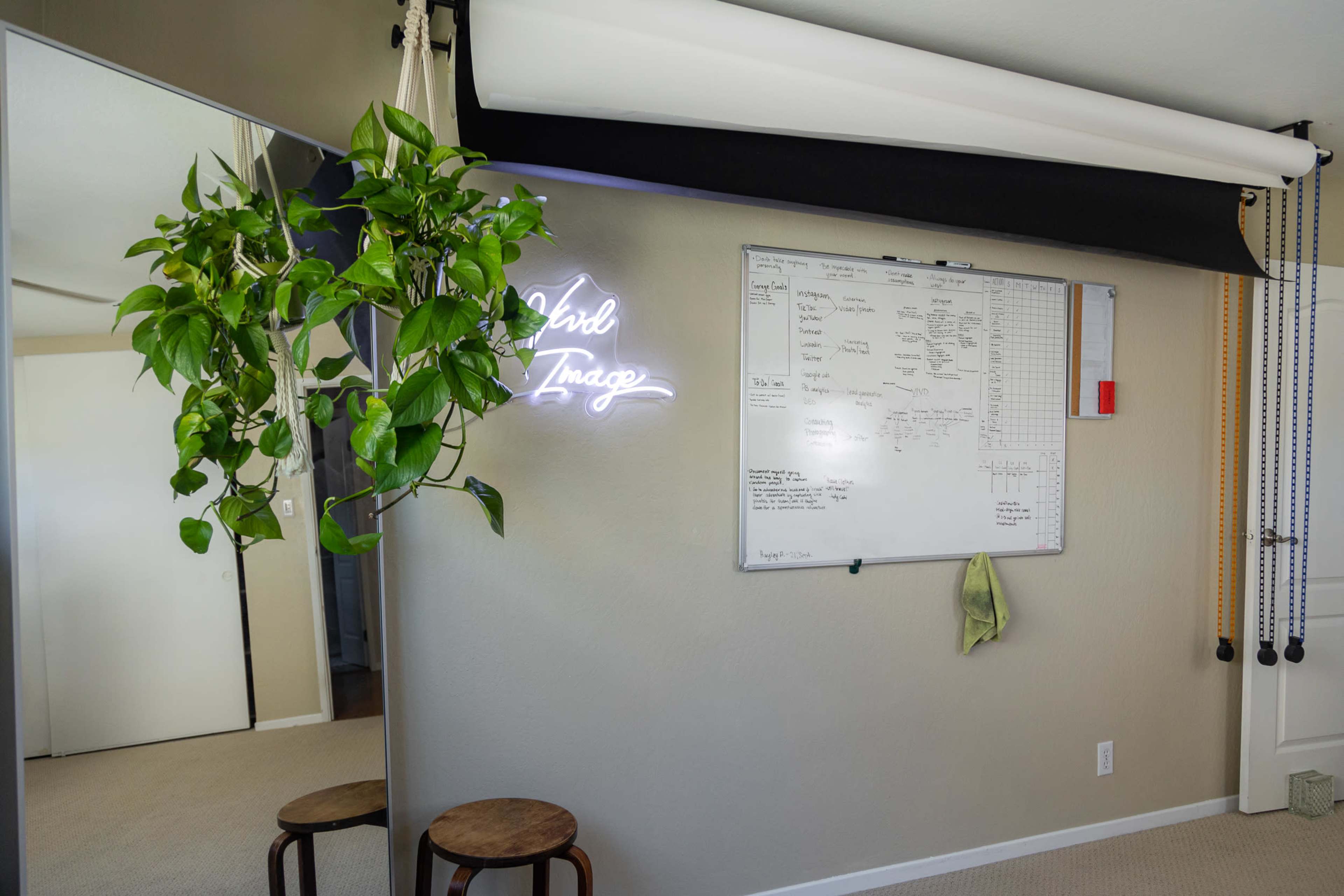 A room featuring a neon sign that says "Girl Image," a whiteboard filled with notes, a hanging plant, and two wooden stools.