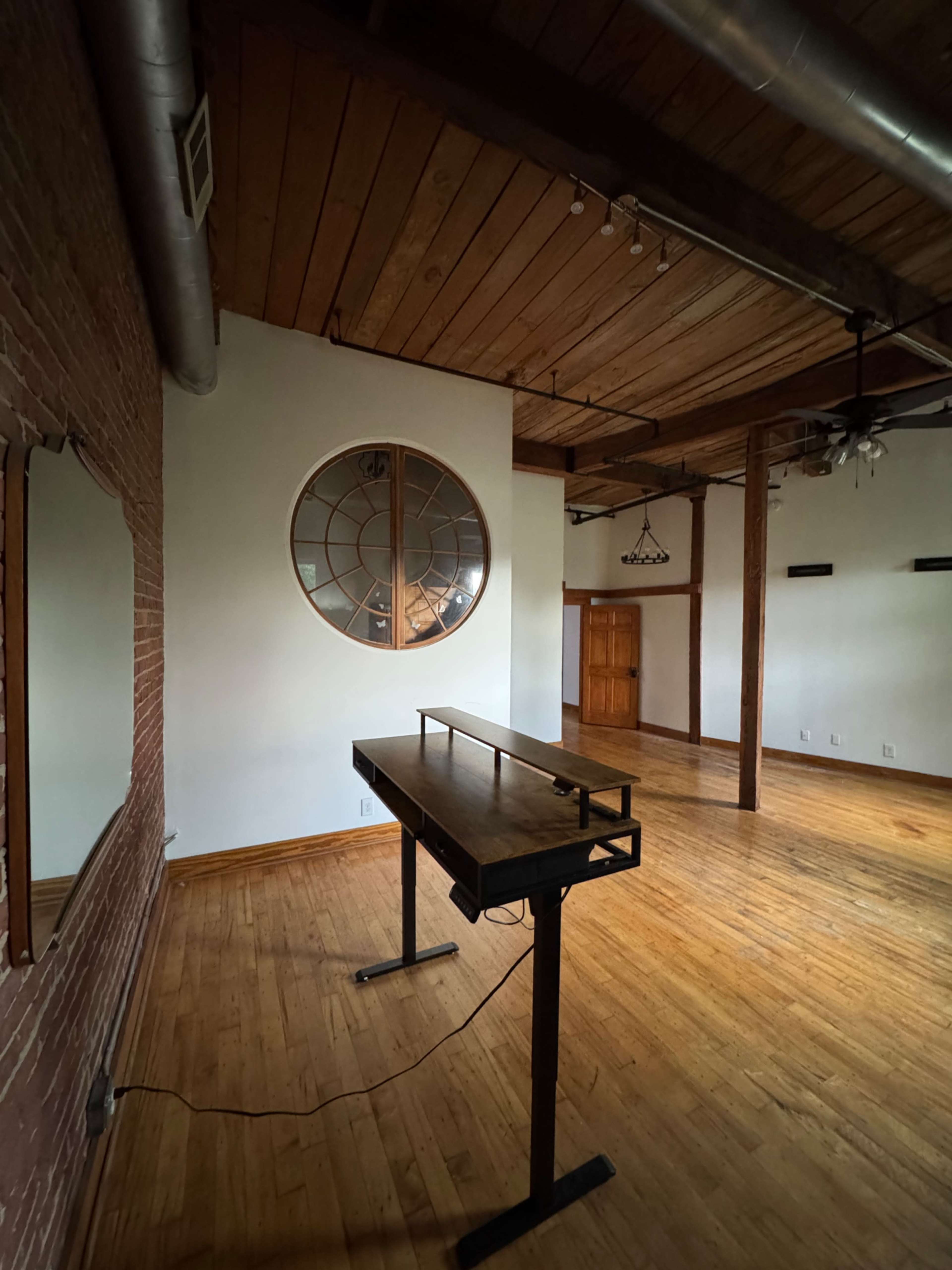 The image shows an empty room with wooden floors, exposed brick walls, and a large circular window, featuring a standing desk in the center.