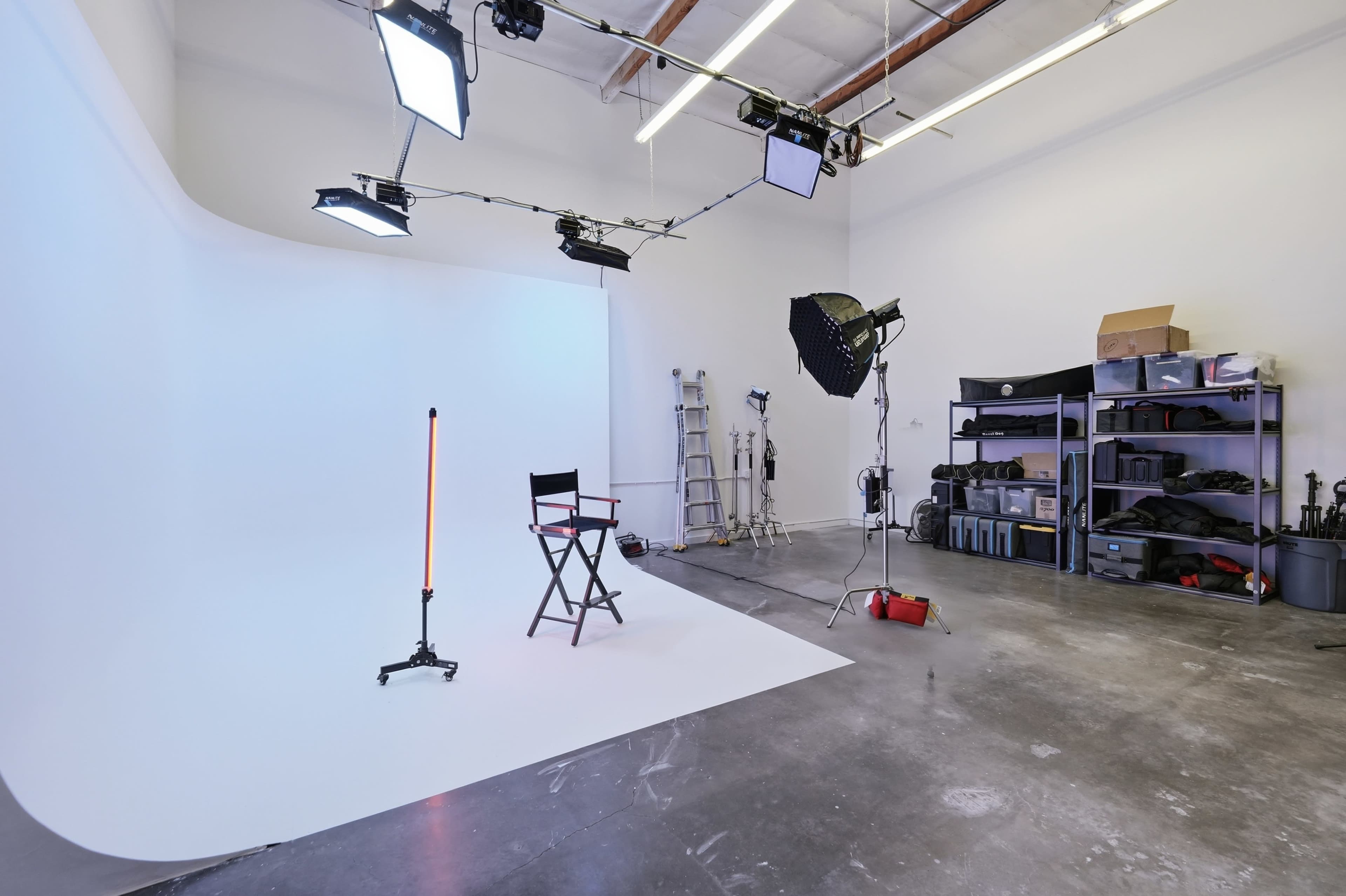 The image shows a photography studio featuring a white backdrop, a director's chair, various lighting equipment, and storage shelves filled with gear.