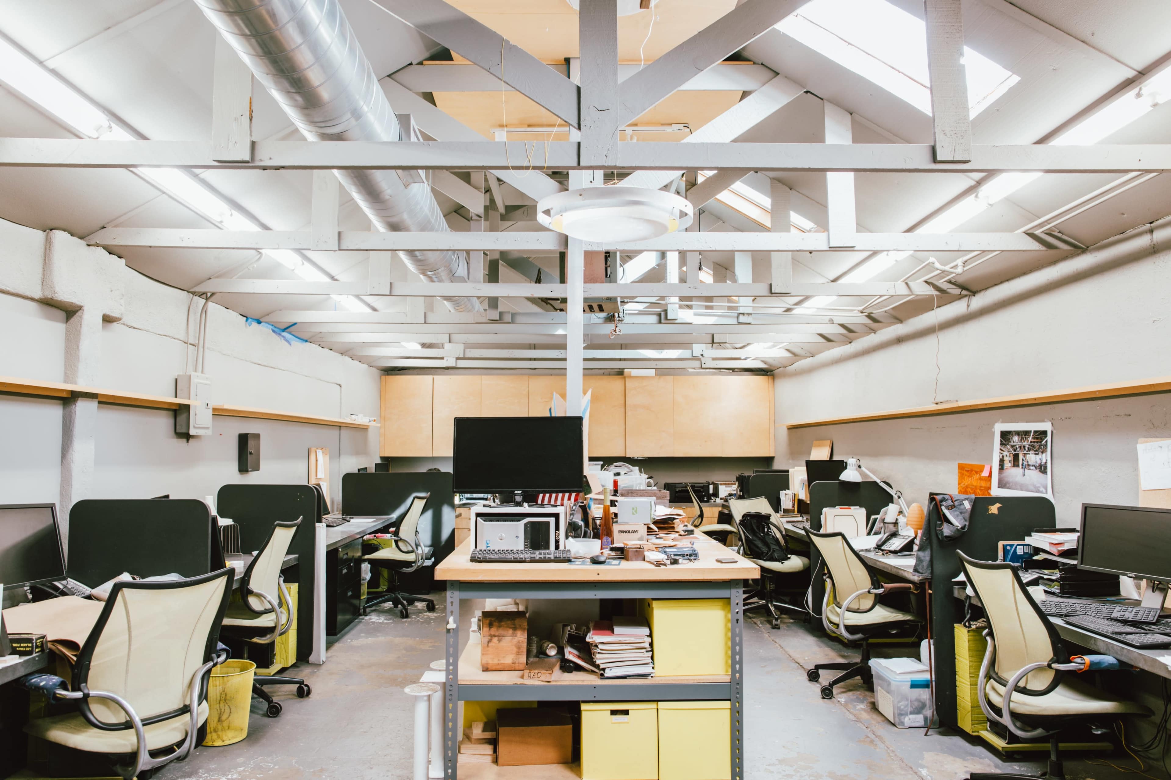 An open office space with multiple workstations, each equipped with computers and office supplies, under a vaulted ceiling with exposed beams.