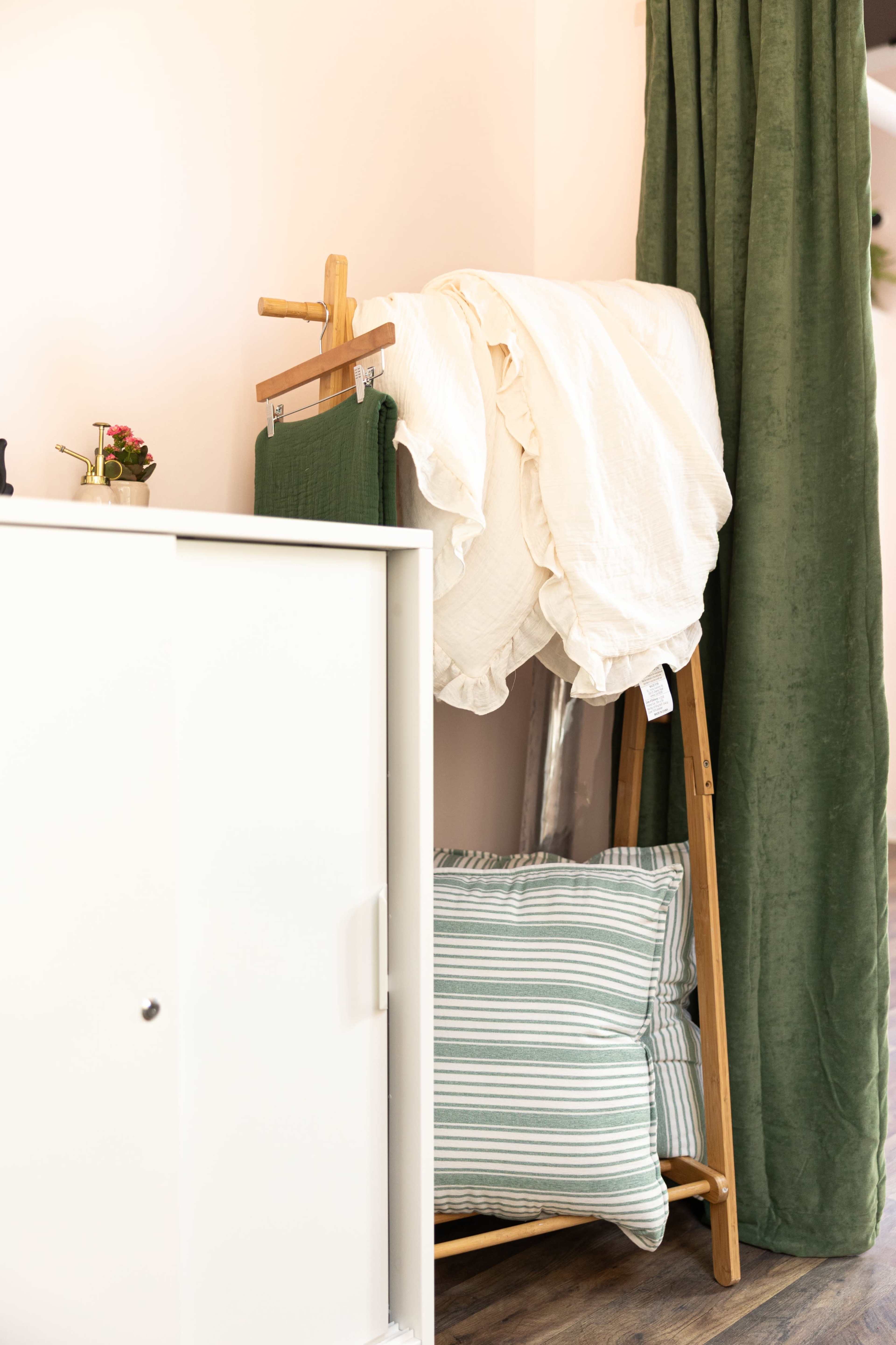 A white cabinet stands next to a wooden rack holding a white blanket and a green fabric, with striped pillows placed nearby on the floor.