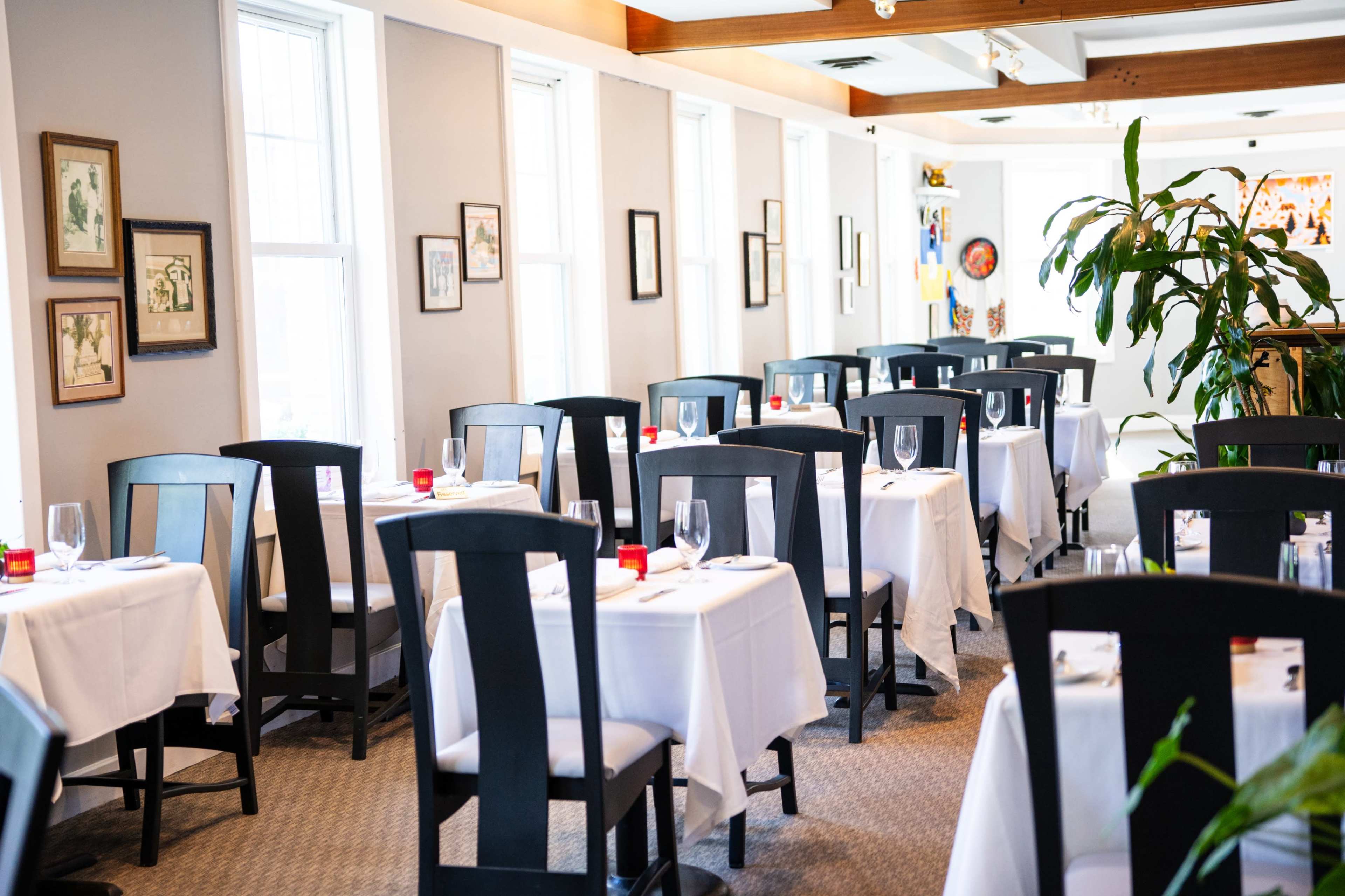 The image shows a dining room with several tables covered in white tablecloths, surrounded by black chairs and adorned with glassware.