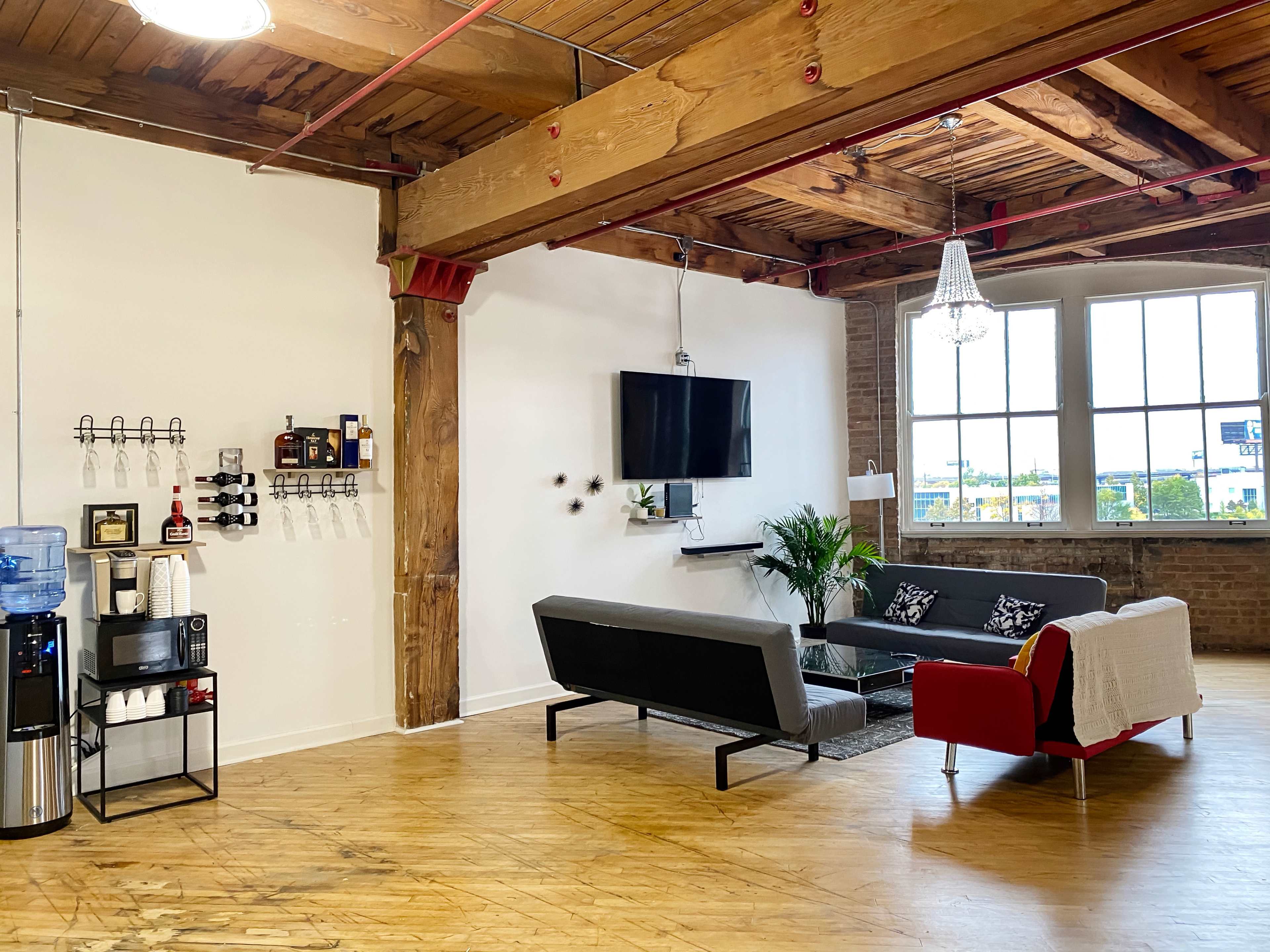 The image shows a modern office lounge area featuring a gray and red sofa, a glass coffee table, a television mounted on the wall, and a water cooler beside a coffee station under exposed wooden beams and large windows.