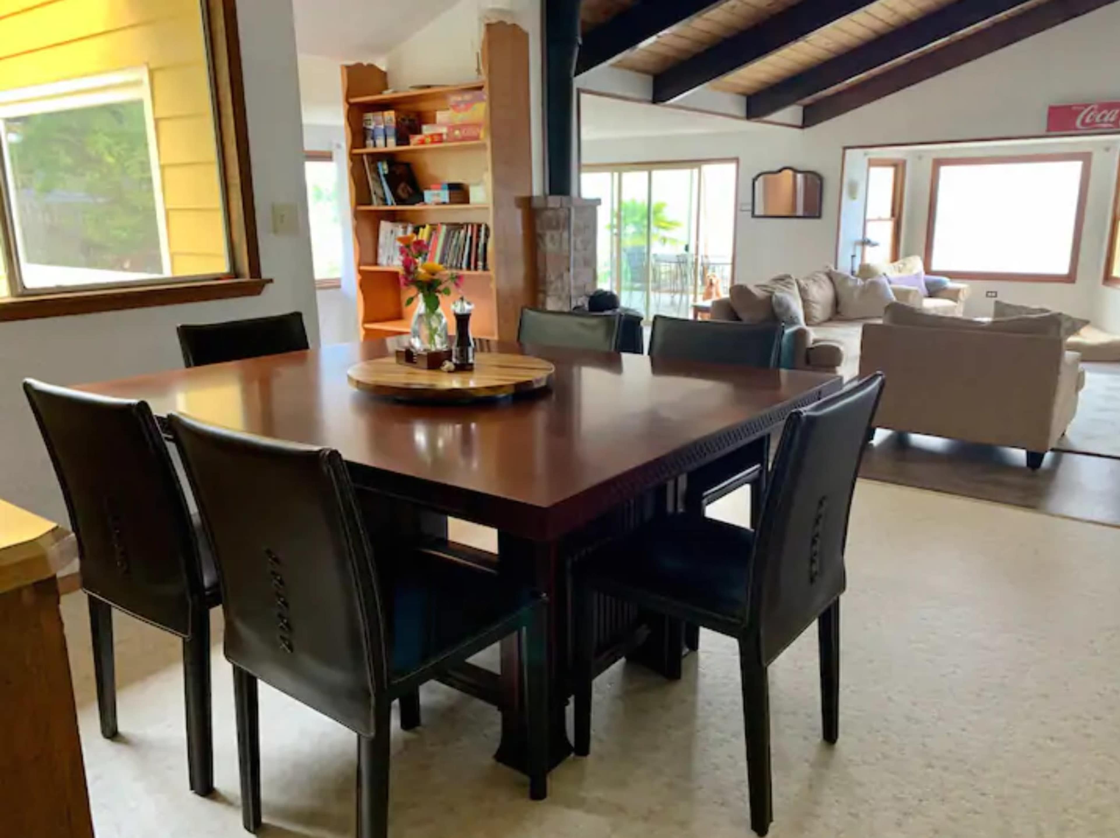 A wooden dining table with black chairs is positioned in a bright room that connects to a living area with a couch and large windows.