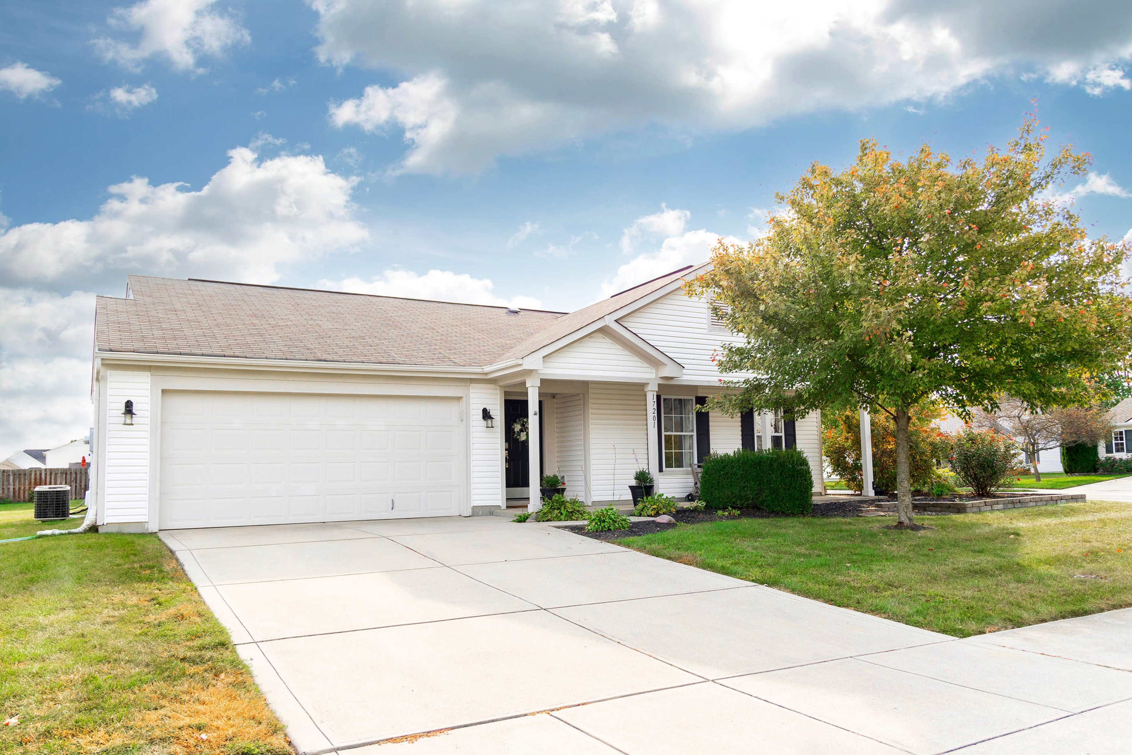A single-story house with a front porch and a two-car garage is set on a neatly paved driveway, surrounded by grass and a tree.