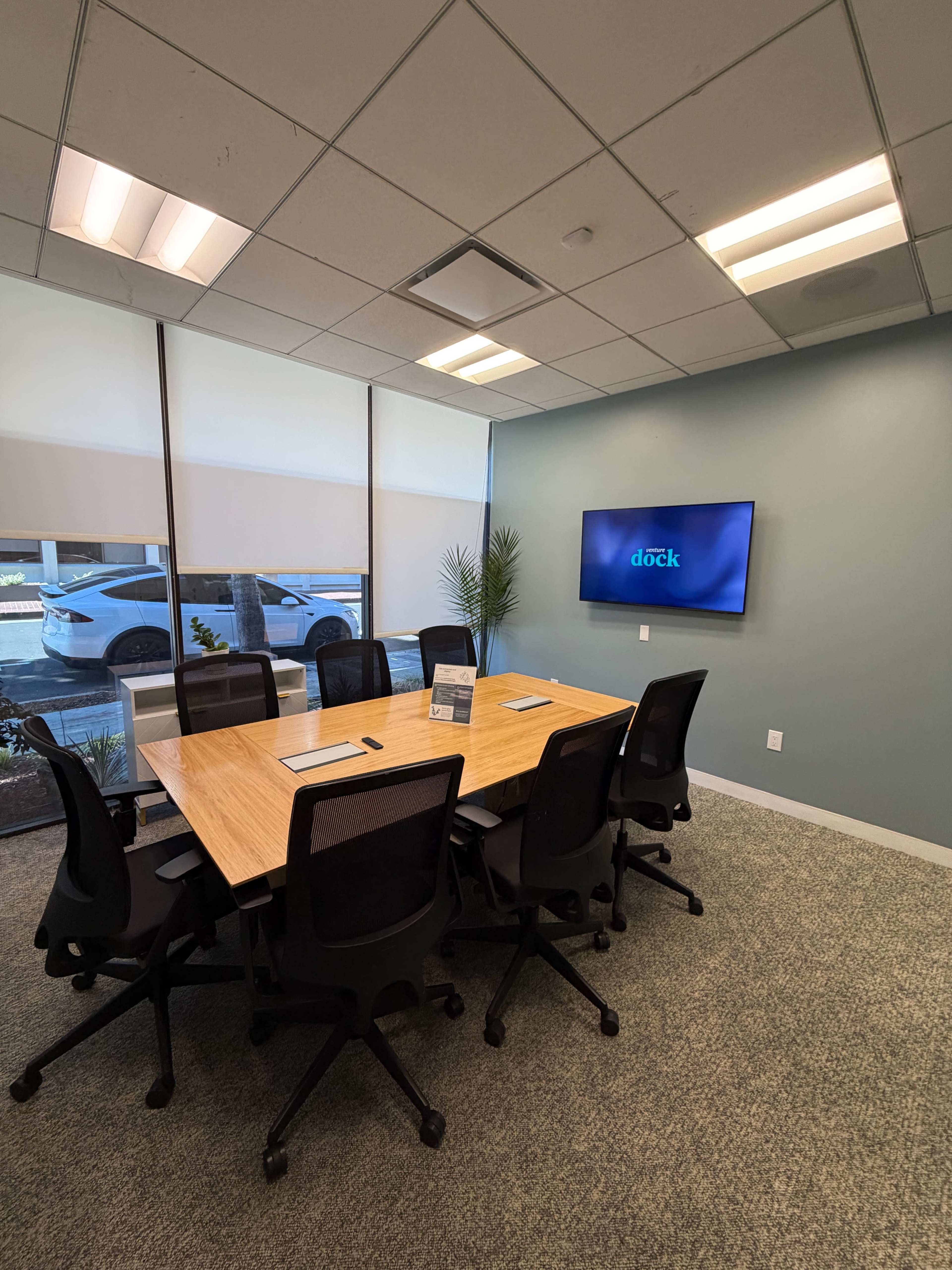 The image shows a conference room with a large wooden table, six black chairs, a TV mounted on the wall, and windows that allow natural light to enter.
