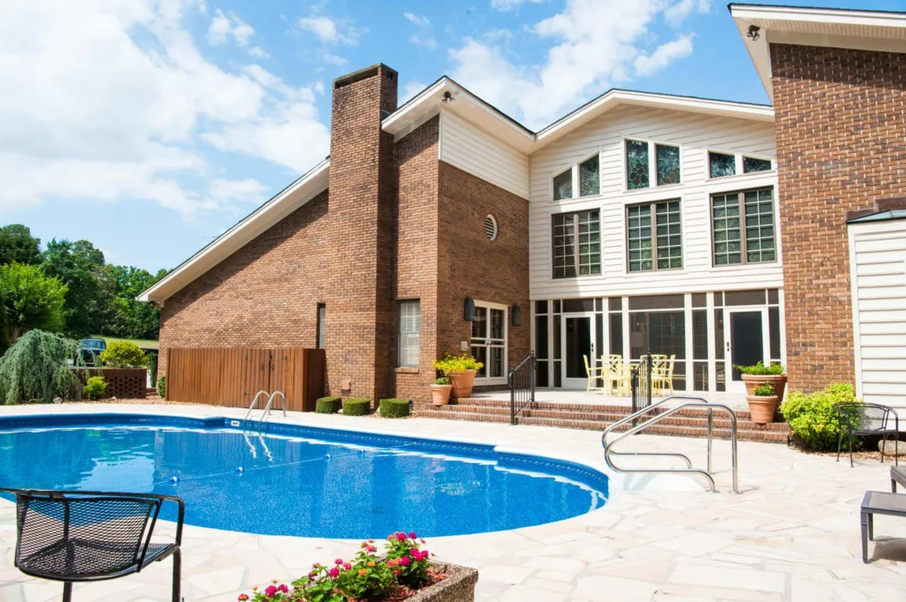 A brick house with large windows overlooks a blue swimming pool surrounded by stone patio and greenery.