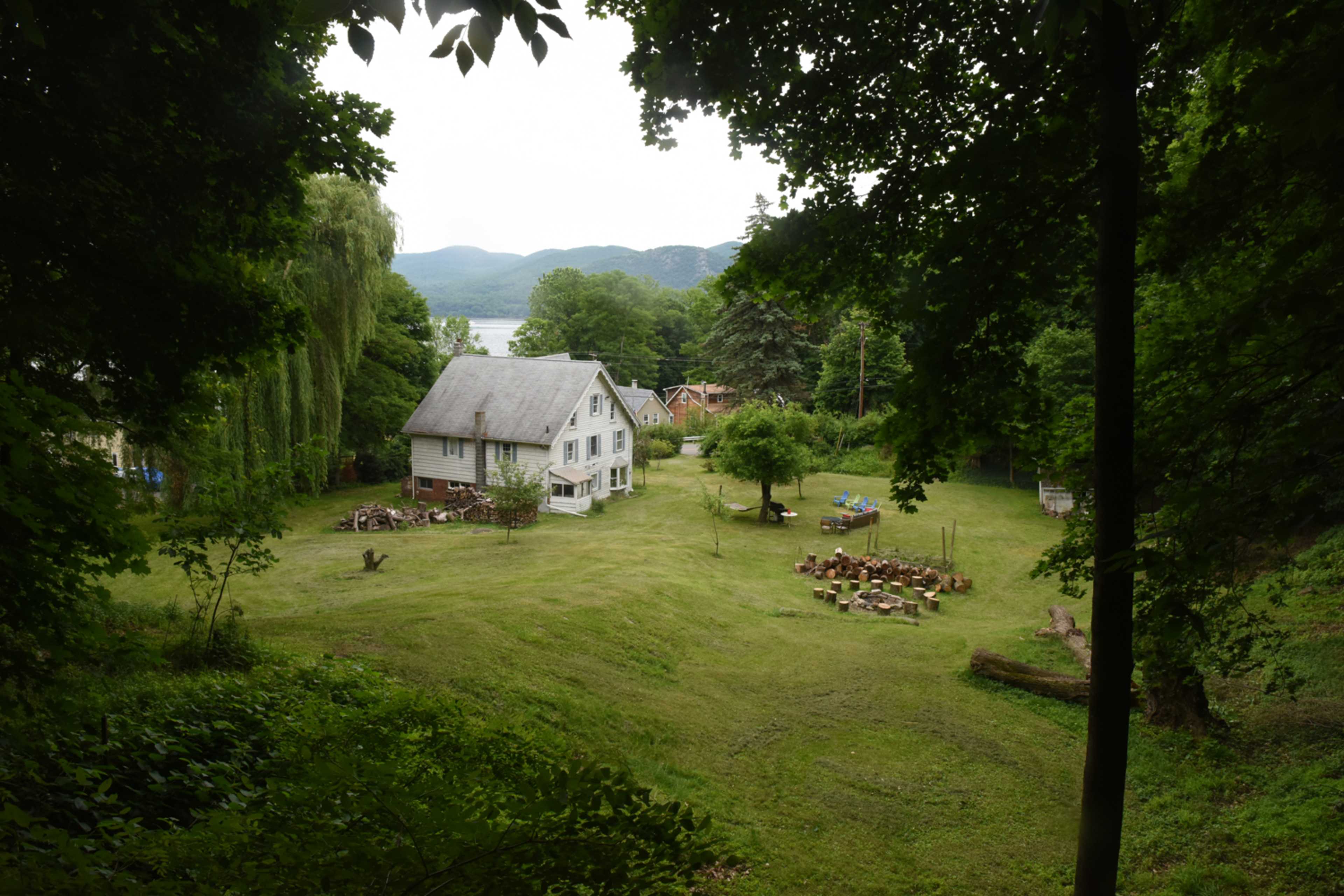A white two-story house sits in a grassy clearing surrounded by trees, with outdoor seating and a firewood stack nearby.