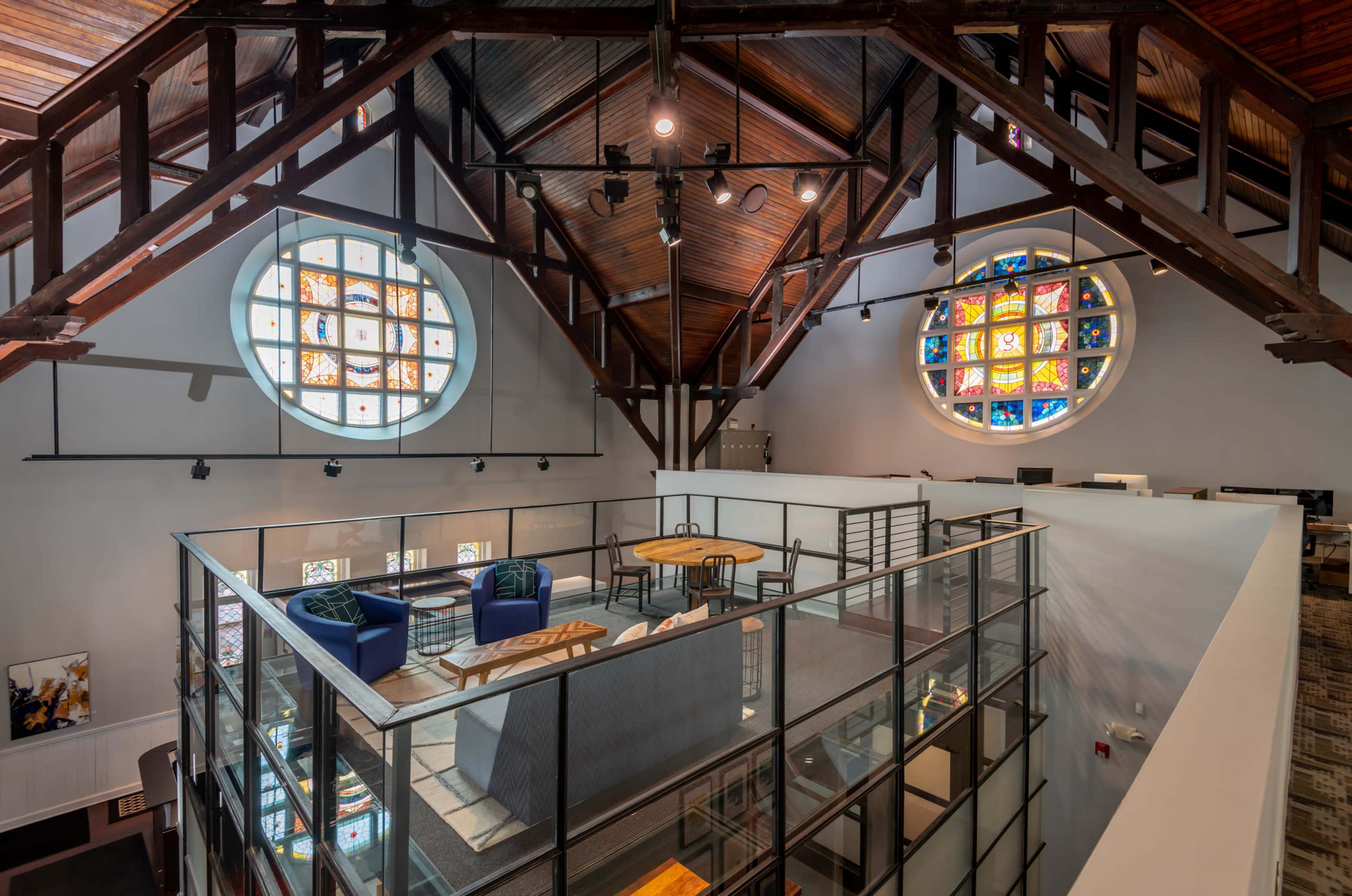 A modern office interior with a glass-walled mezzanine overlooking a space with stained glass windows and exposed wooden beams.