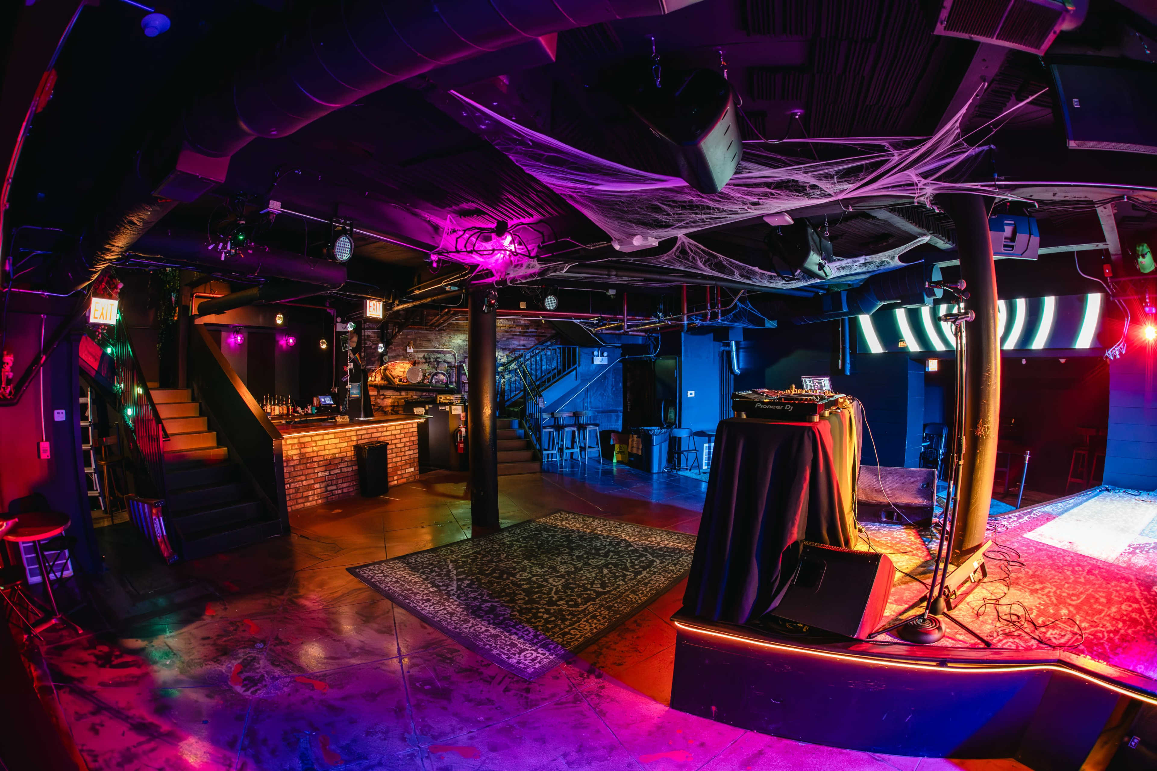 The image shows an empty nightclub interior with colorful lighting, a DJ booth, and a bar area.